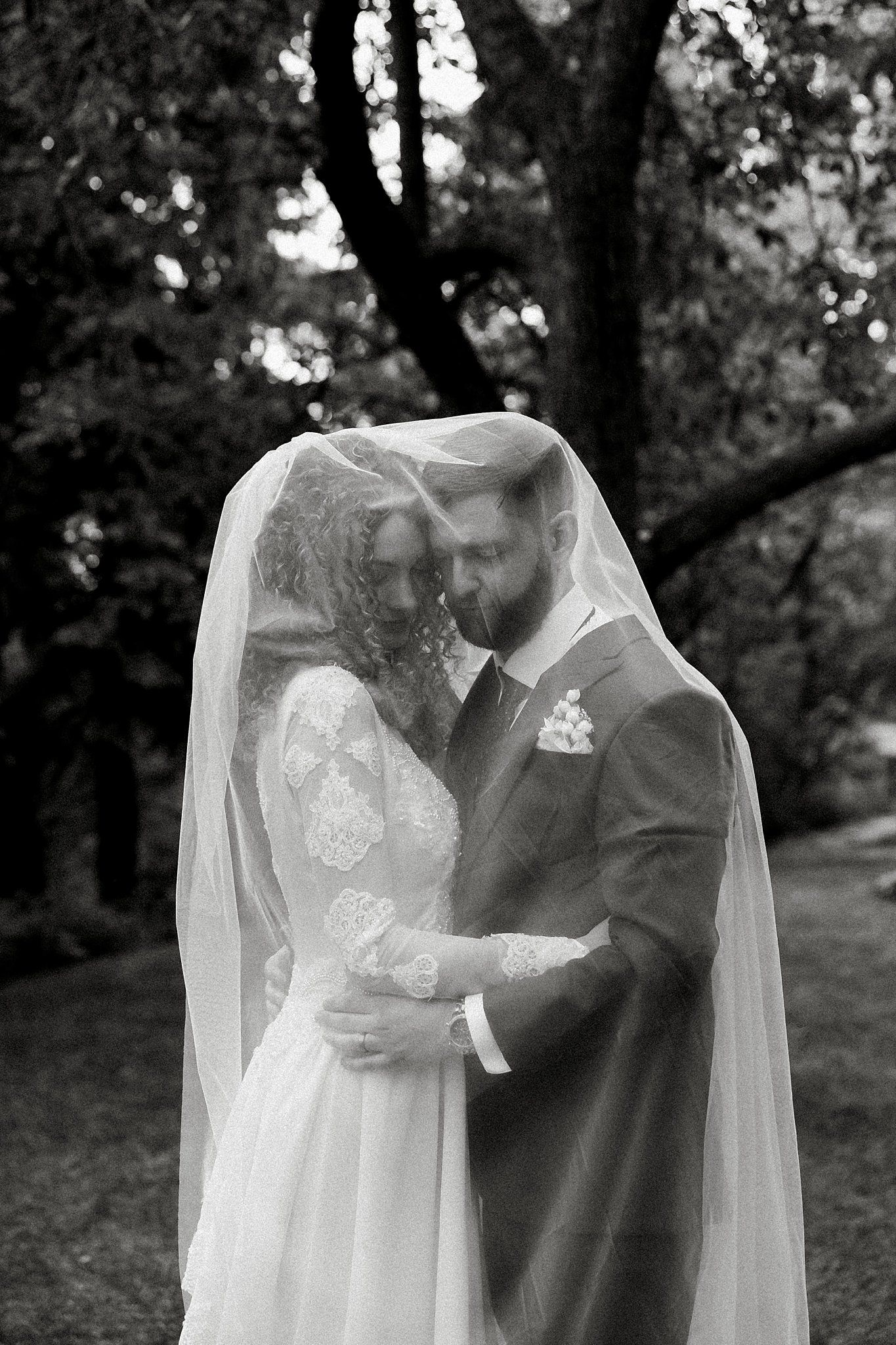 Black and white wedding portrait under trees showing couple embracing beneath a flowing veil.