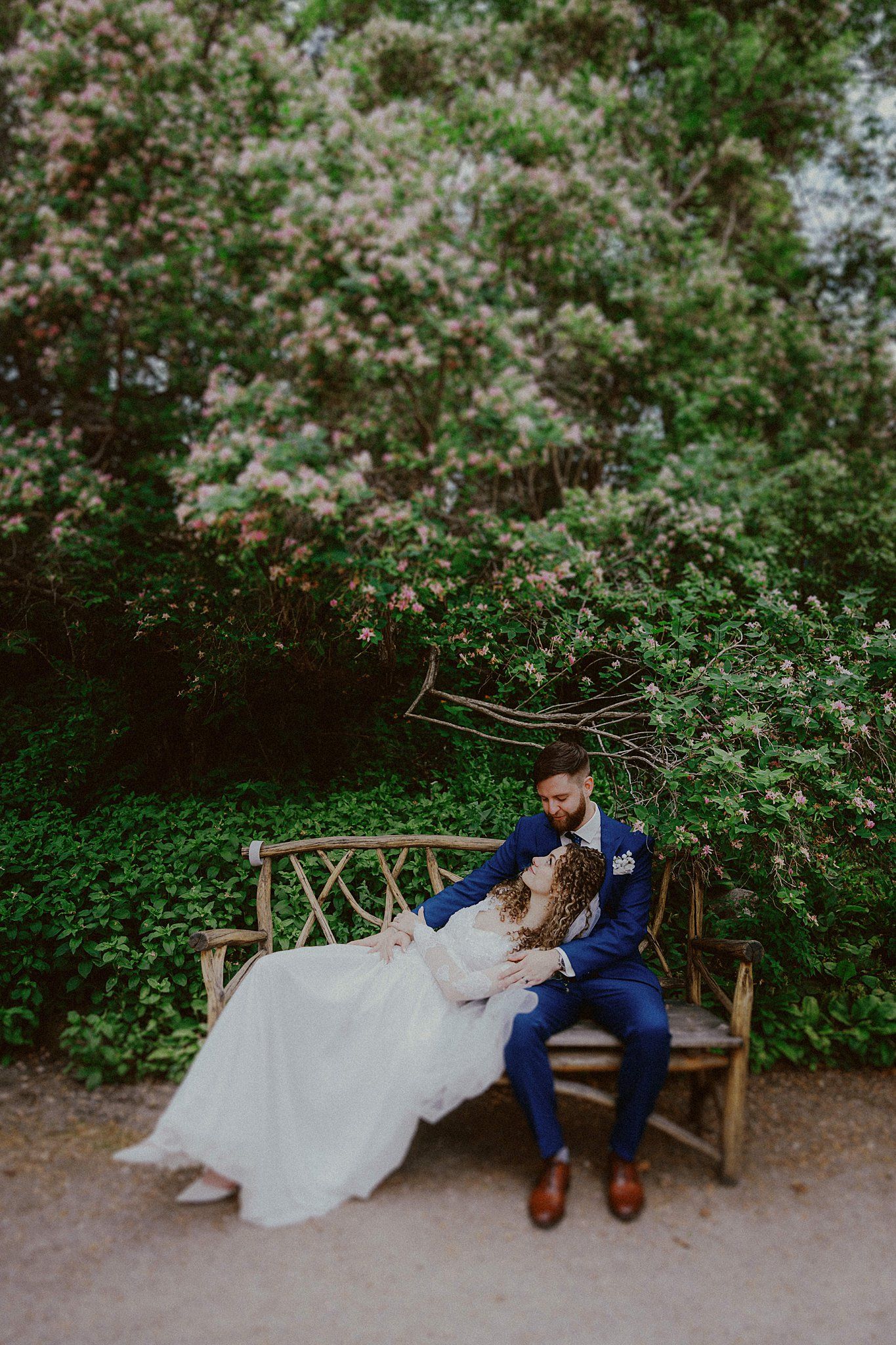 Couple relaxing on a wooden bench surrounded by flowering bushes in a garden setting.