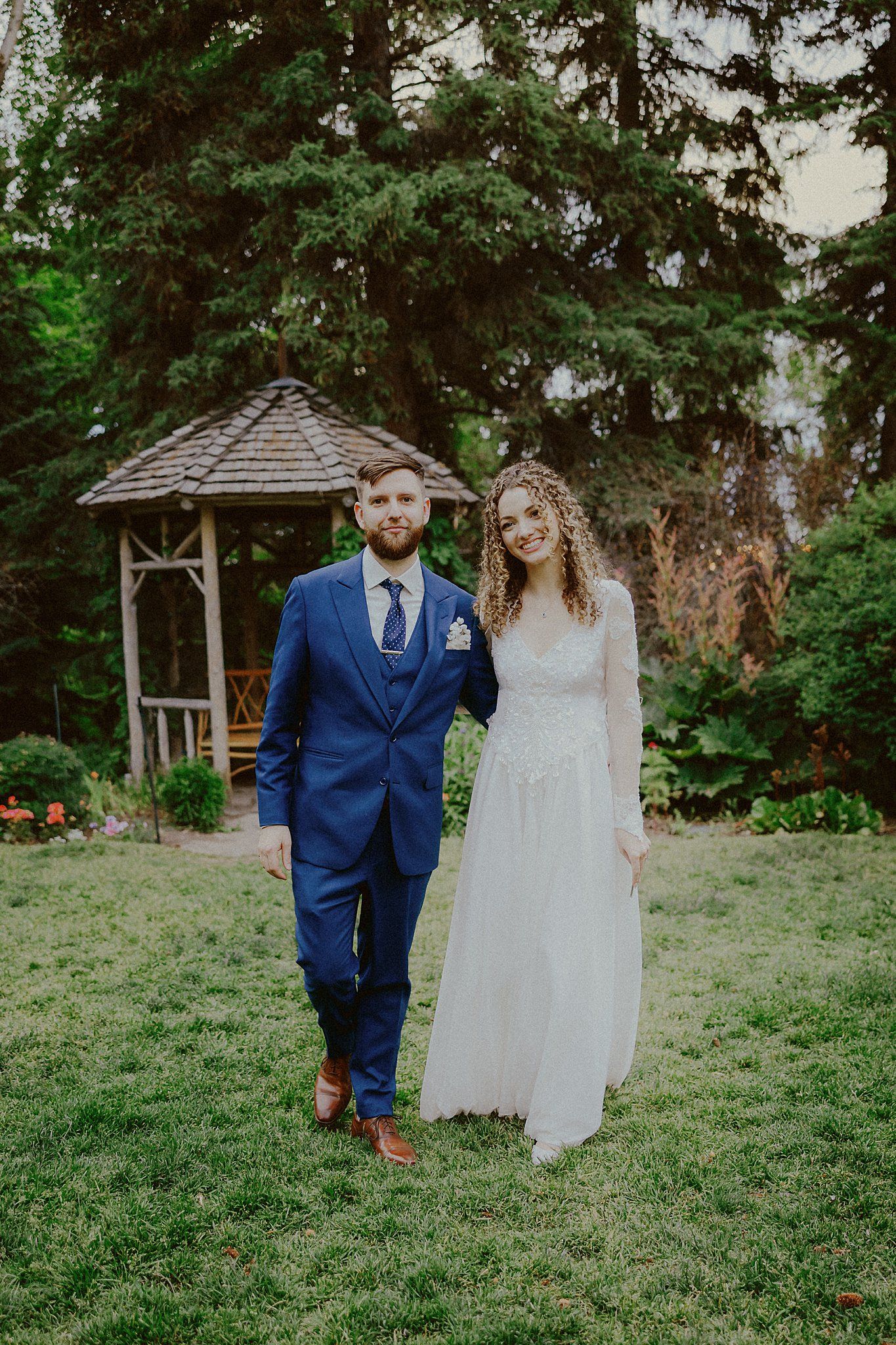 A bride and groom stand together on a green lawn with a wooden gazebo and evergreen trees in the background.