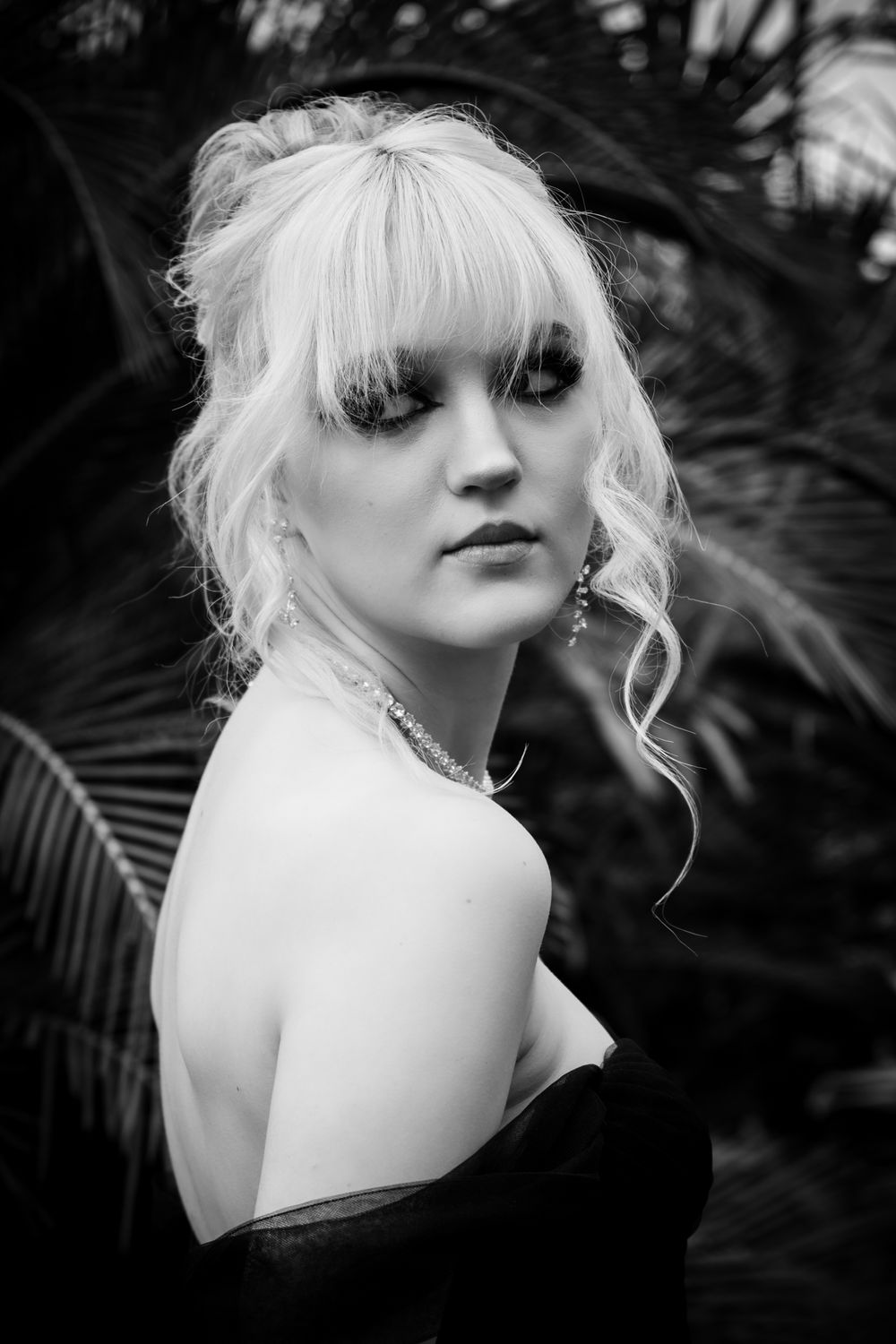 Black and white portrait of a platinum blonde in an off-shoulder dress against a dark palm leaf background.