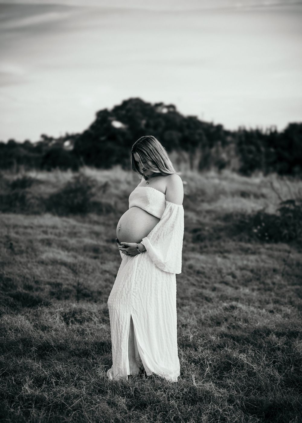 Black and white artistic photograph of a person in a flowing white dress standing on a grassy hillside.