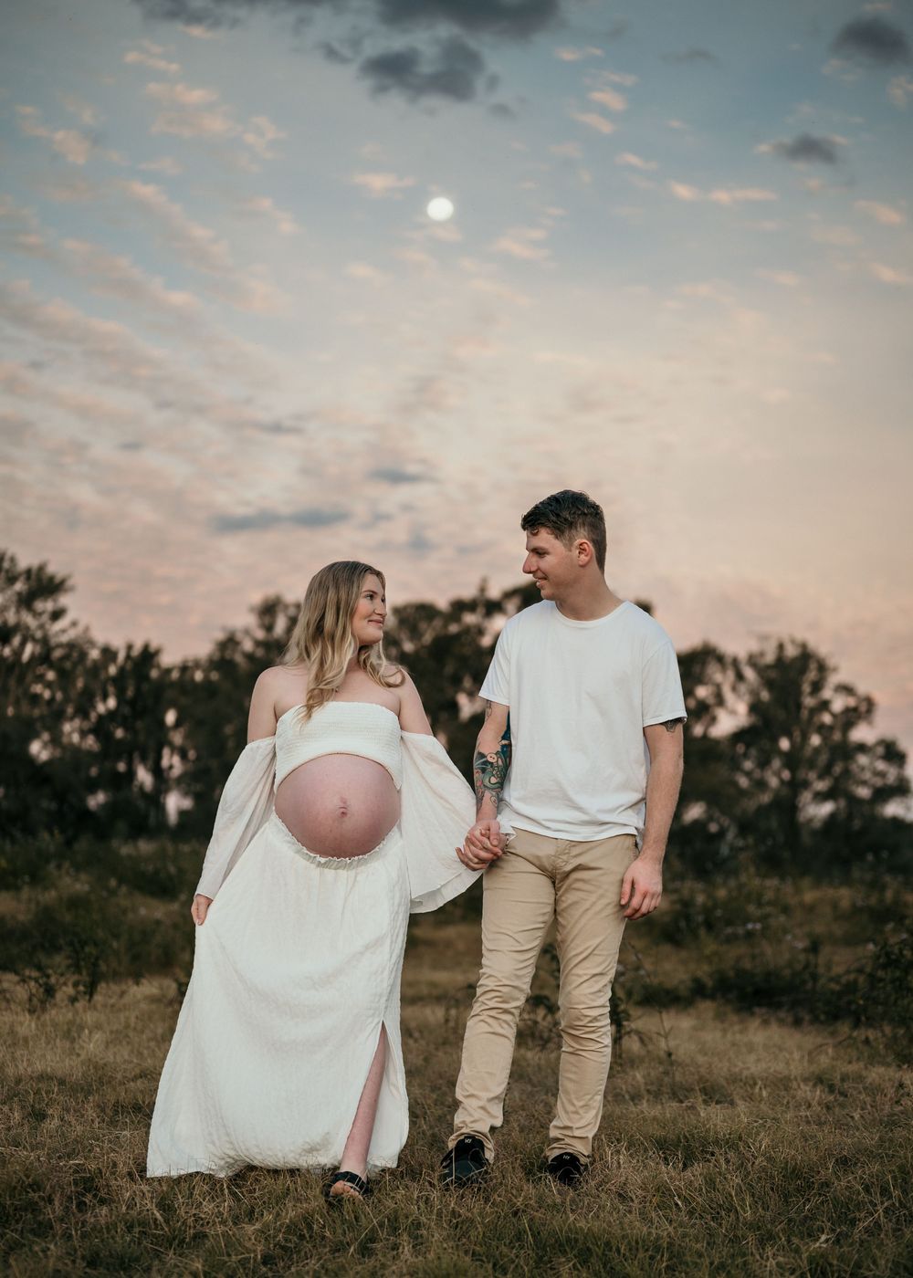 A couple in white and beige outfits walks together in a grassy field at sunset while holding a pink balloon.