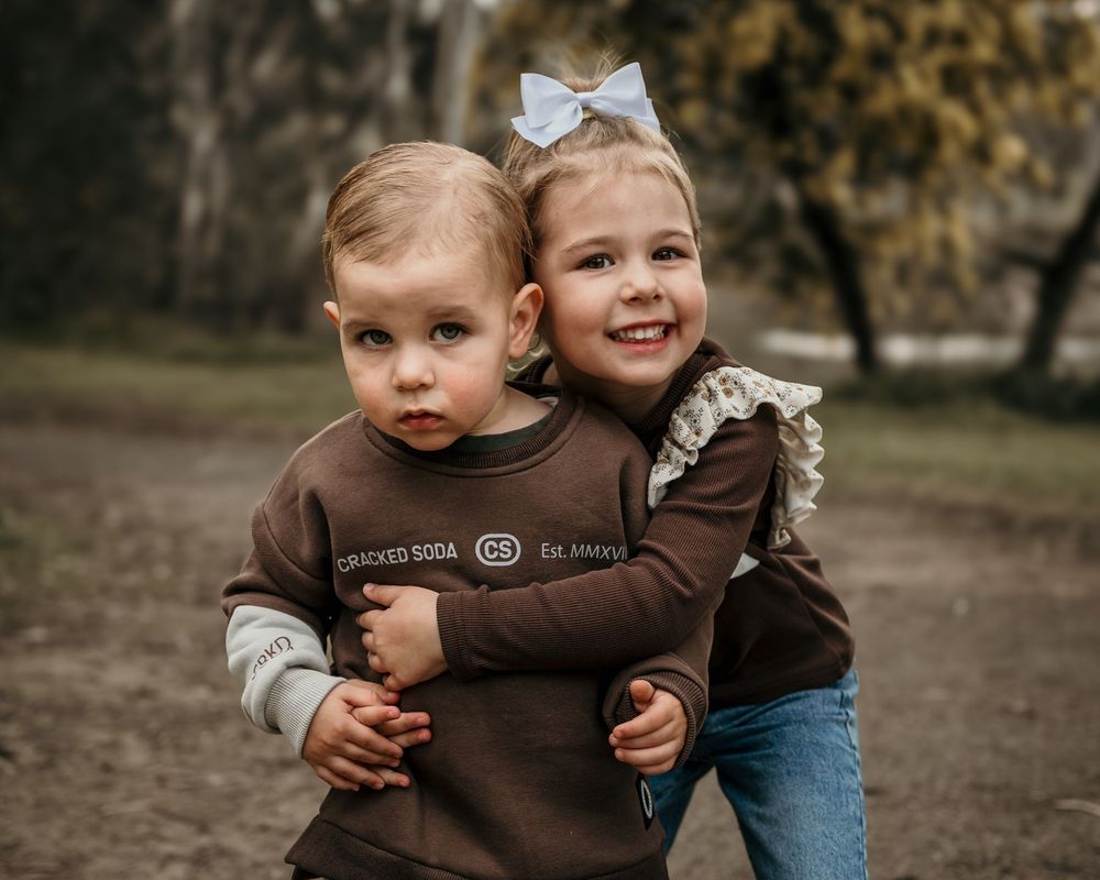 Two children wearing brown and denim outfits play and hug while laughing outdoors in an autumn setting.