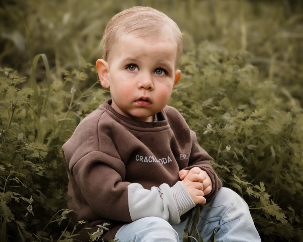 A child in a brown hoodie sits thoughtfully in a grassy outdoor setting at dusk.