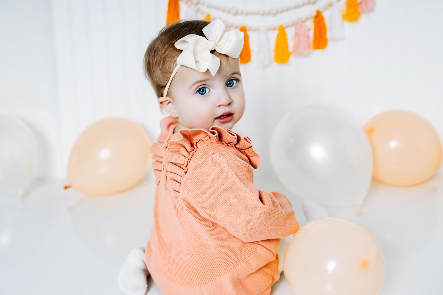 Baby in peach-colored ruffled outfit poses with orange tassels and peach balloons at party photoshoot.