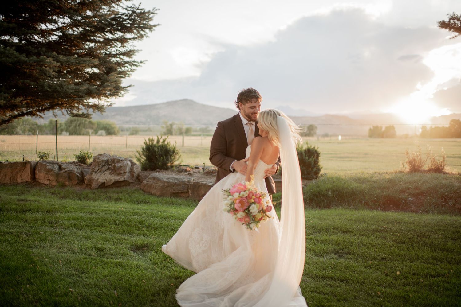 Romantic wedding couple embraces in a sunlit meadow with mountains and evergreen trees in the background at sunset.