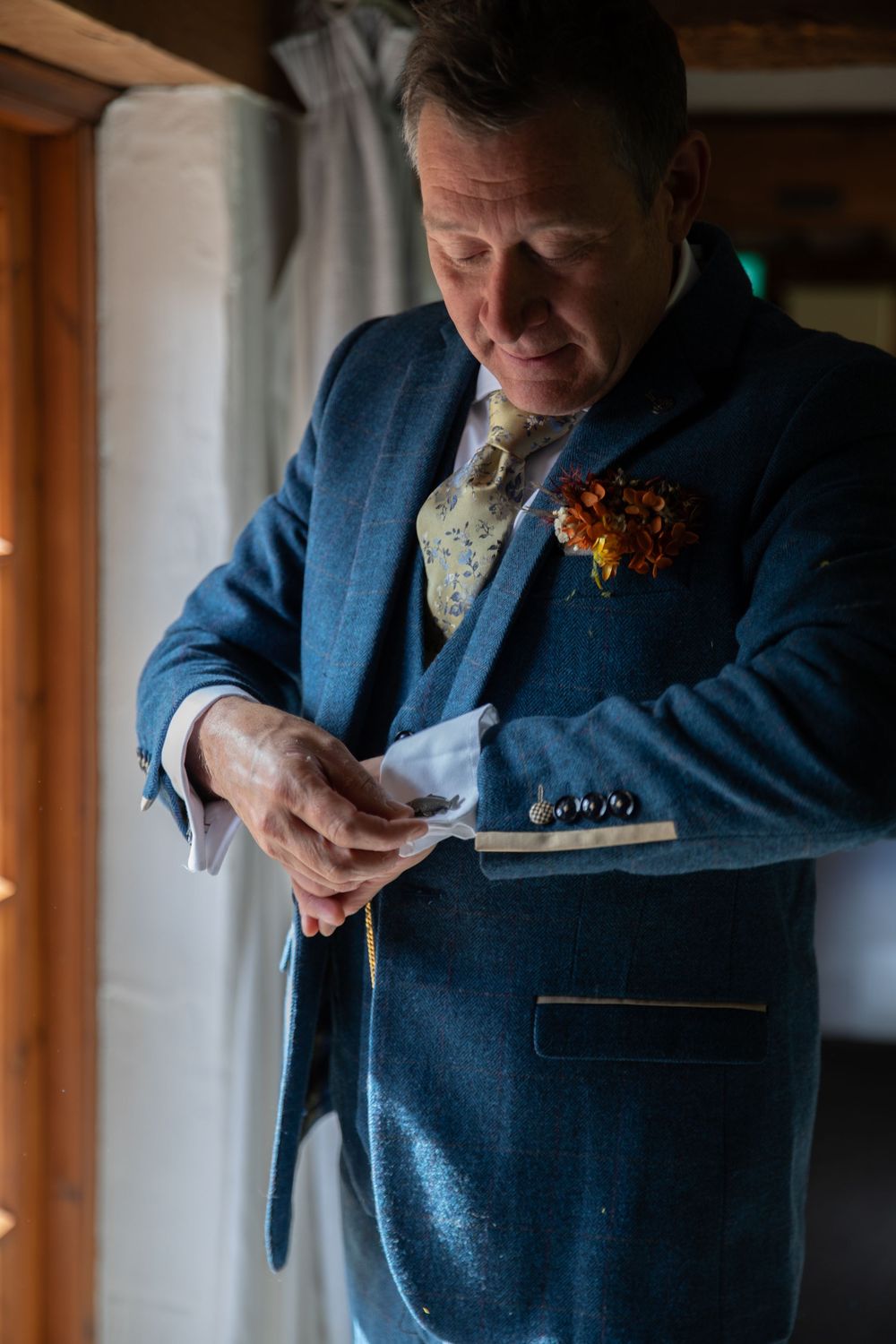A person in a blue suit jacket with a floral boutonniere adjusts their cuffs in natural window light.