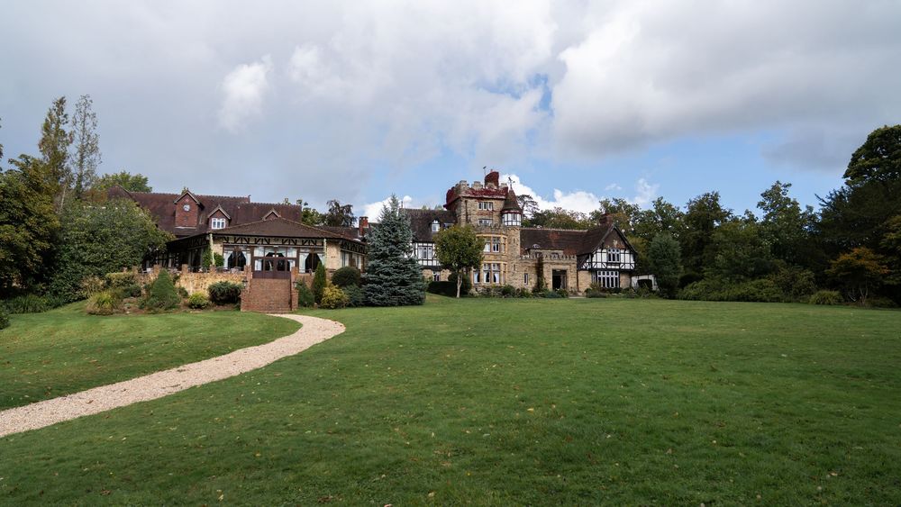 A panoramic view shows a historic stone mansion estate with manicured lawns and gardens stretching across rolling countryside.