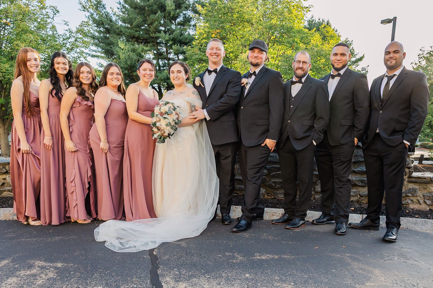 Wedding party in pink bridesmaid dresses and black tuxedos pose together outdoors on a sunny day.
