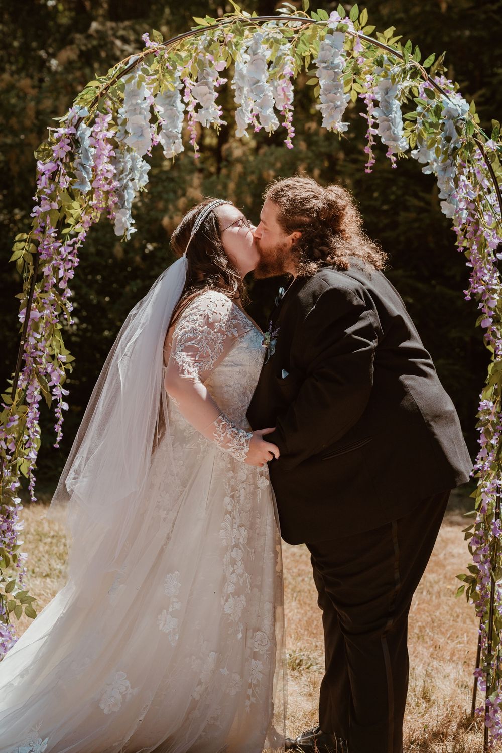 Bride and groom share first kiss under a floral arch adorned with purple and white wisteria.