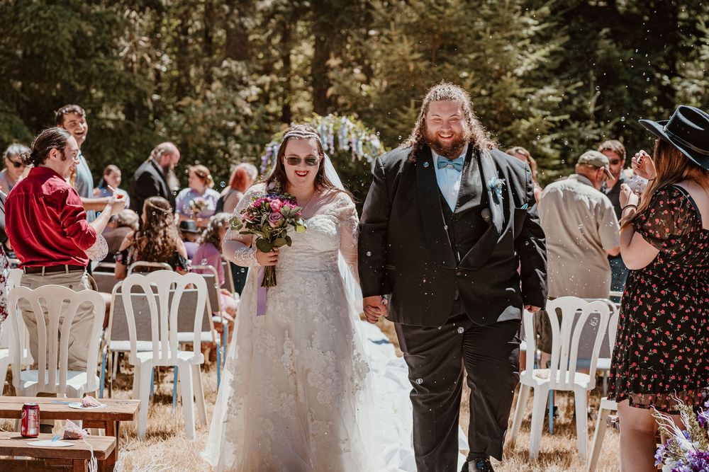 Smiling newlyweds walk hand-in-hand down the aisle at an outdoor forest wedding ceremony.