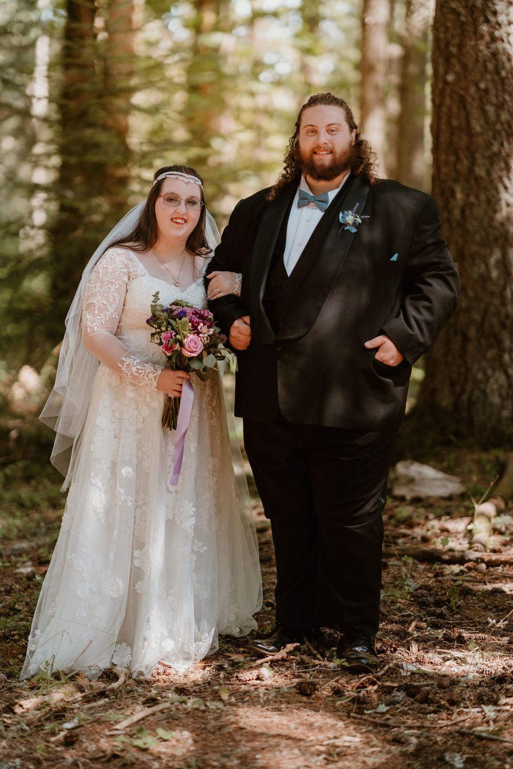 Bride and groom pose together in a sunlit forest, she holds a colorful bridal bouquet.