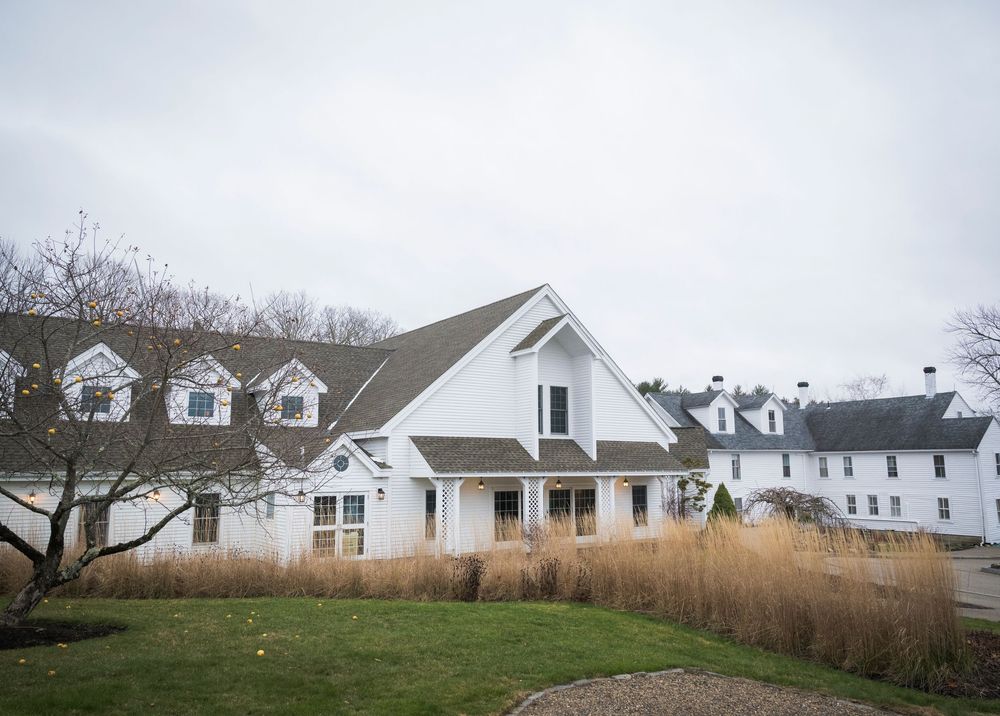 White farmhouse style venue with dormers and rustic exterior surrounded by tall grass on an overcast day.