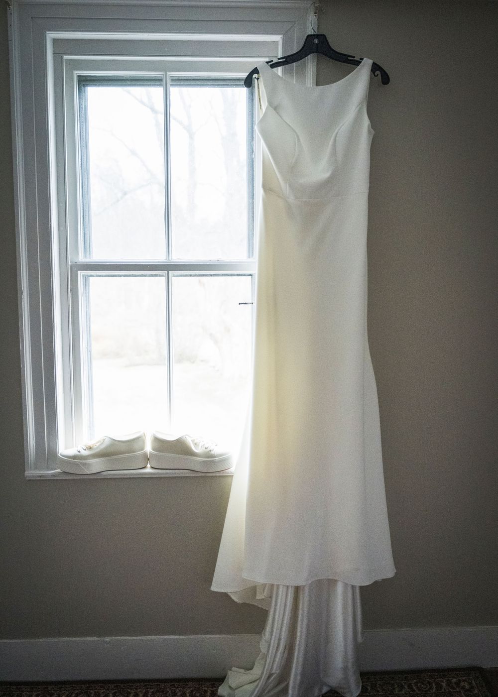 A white wedding dress hangs elegantly by a window, showcasing its simple and classic silhouette.