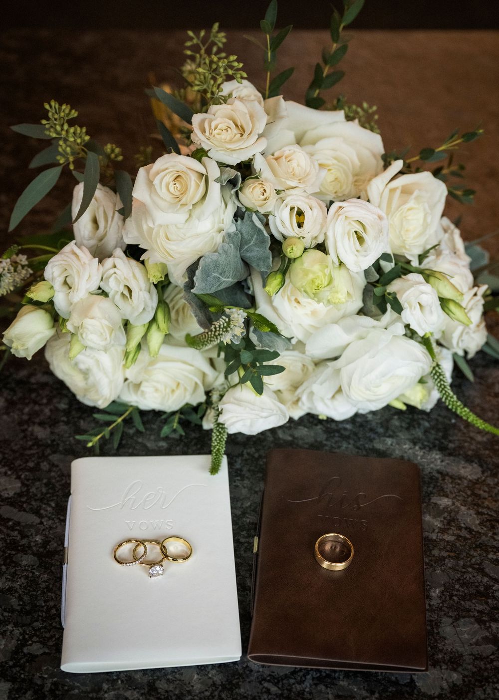 Wedding rings rest on white and brown leather portfolios alongside elegant white rose floral arrangements.