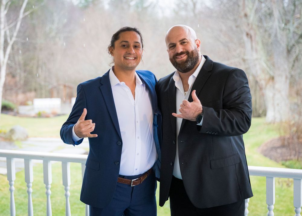 Two businessmen in dark suits and white shirts stand together outdoors giving thumbs up gestures.