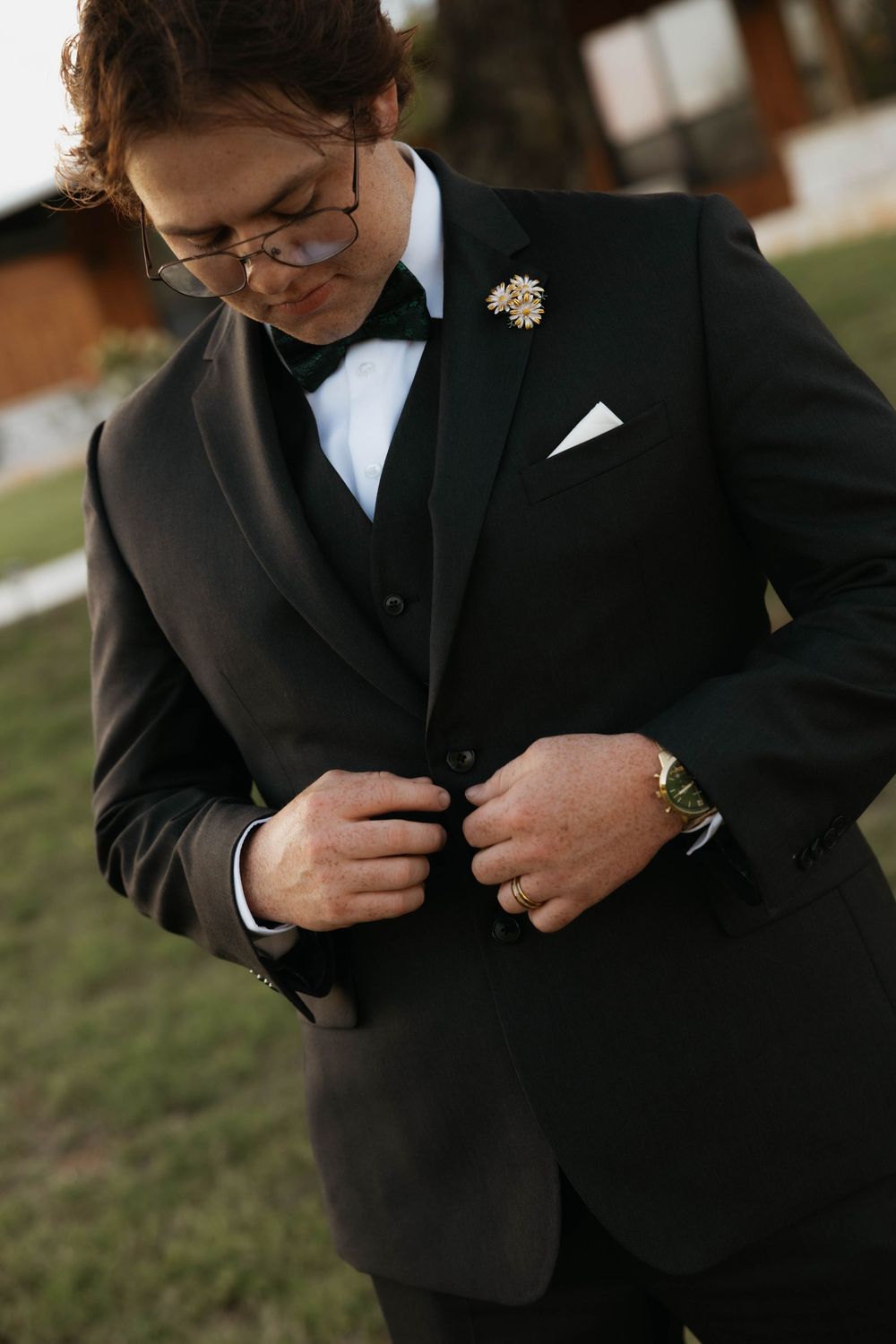 Dapper groom in a black tuxedo with gold boutonniere and watch buttoning his jacket outdoors.