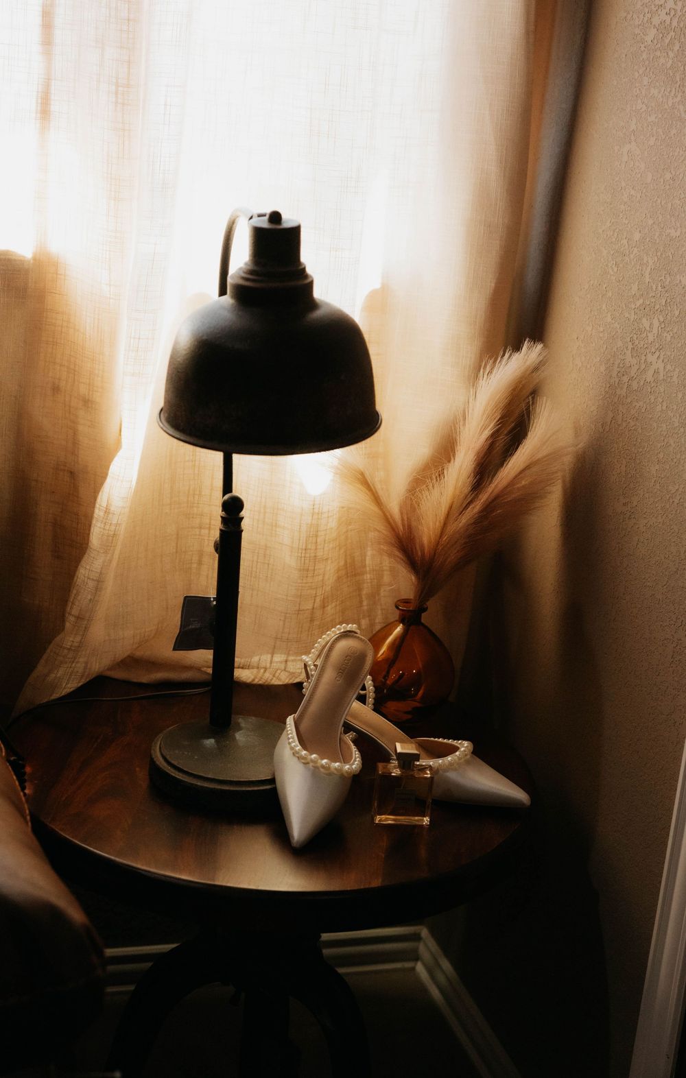 Pearl-trimmed bridal heels and perfume bottle styled on side table with vintage lamp by window.