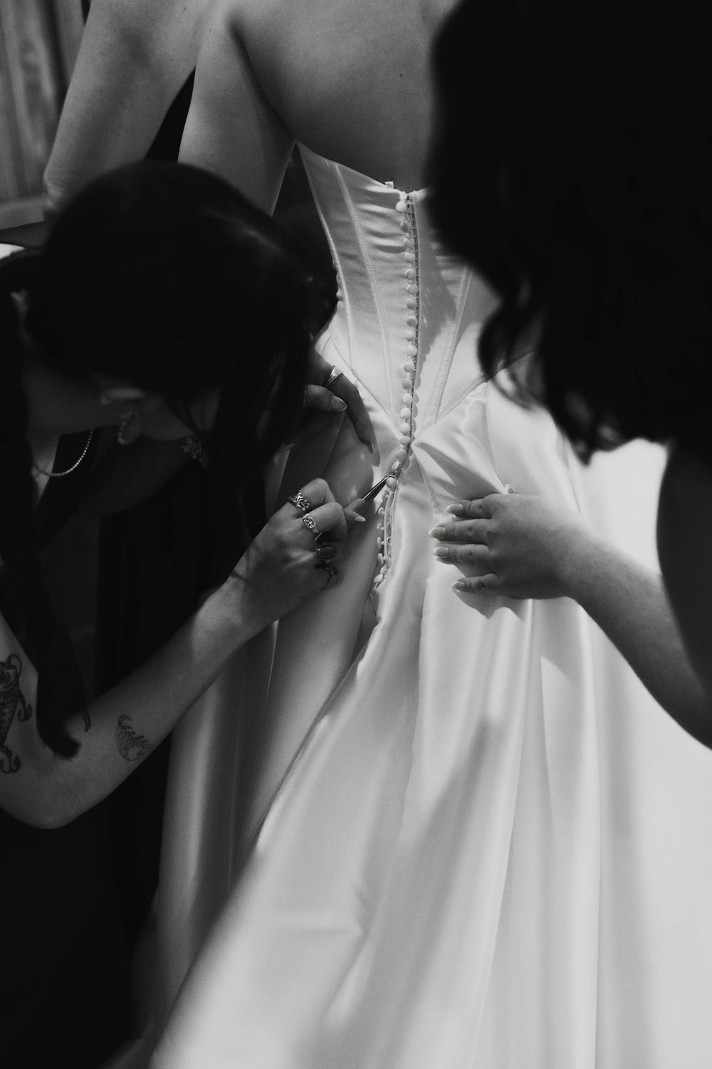 Black and white close-up of bridesmaids fastening pearl buttons on the back of a wedding gown.