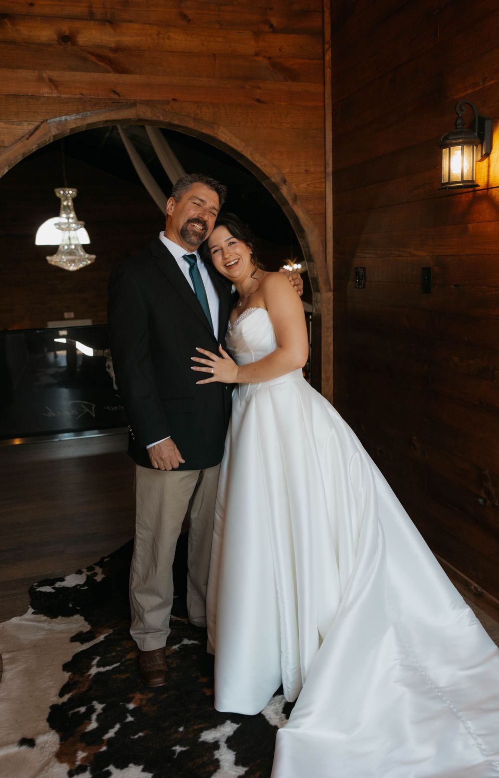 Bride in white ball gown smiles with man in dark suit inside rustic wood-paneled wedding venue with arched doorway.