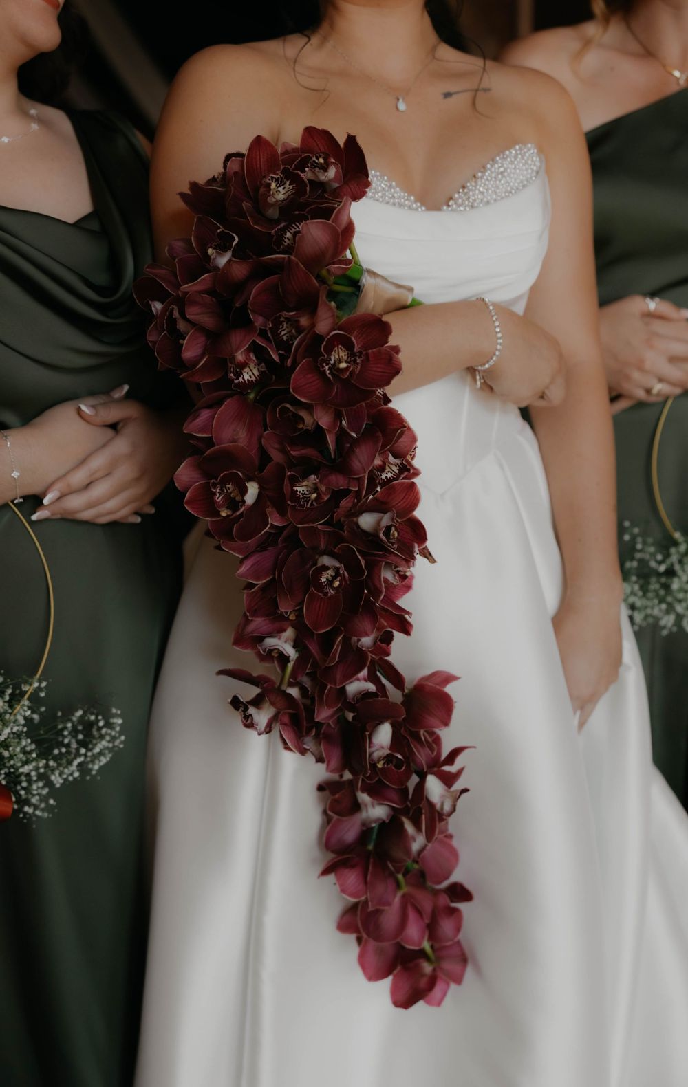 Bride in strapless white gown holds stunning cascading burgundy cymbidium orchid bouquet with bridesmaids in sage green.