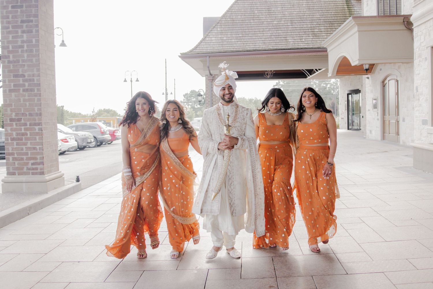 Wedding party in orange floral dresses and white attire poses together in a modern architectural setting.