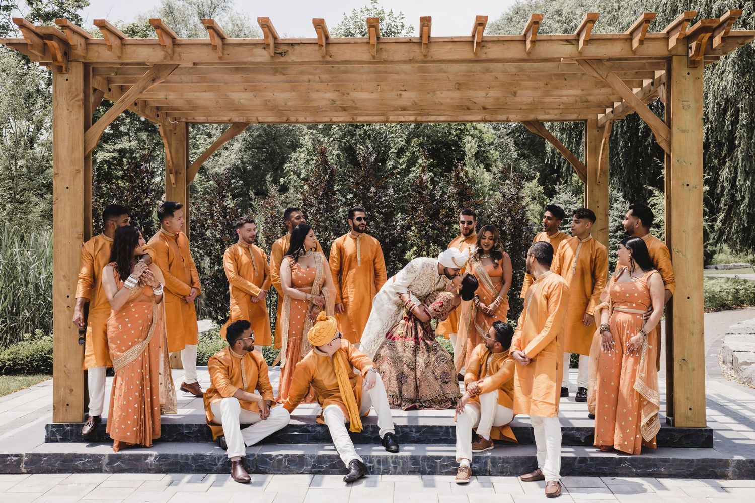 Wedding party in orange and white outfits sitting in a row on outdoor stone steps under a wooden pergola.