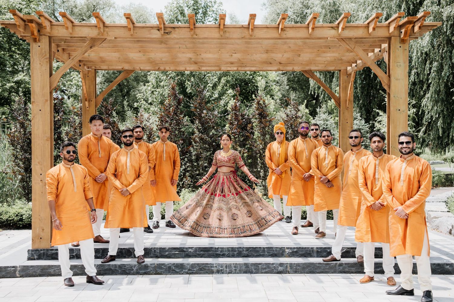 Wedding party members in vibrant orange attire stand beneath a wooden pergola during an outdoor celebration.