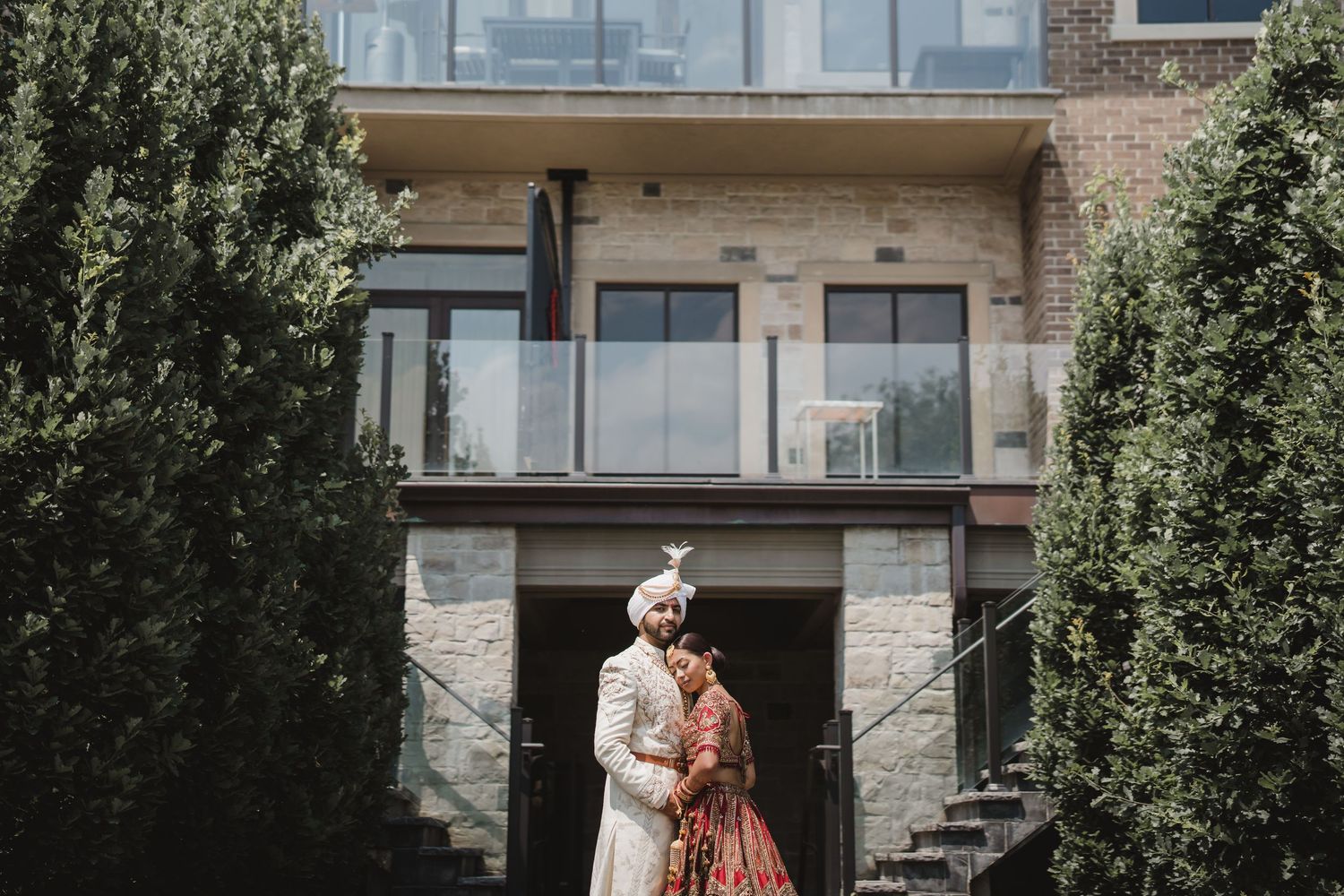 A couple in traditional Indian wedding attire stands together in front of a modern brick building with glass balconies.