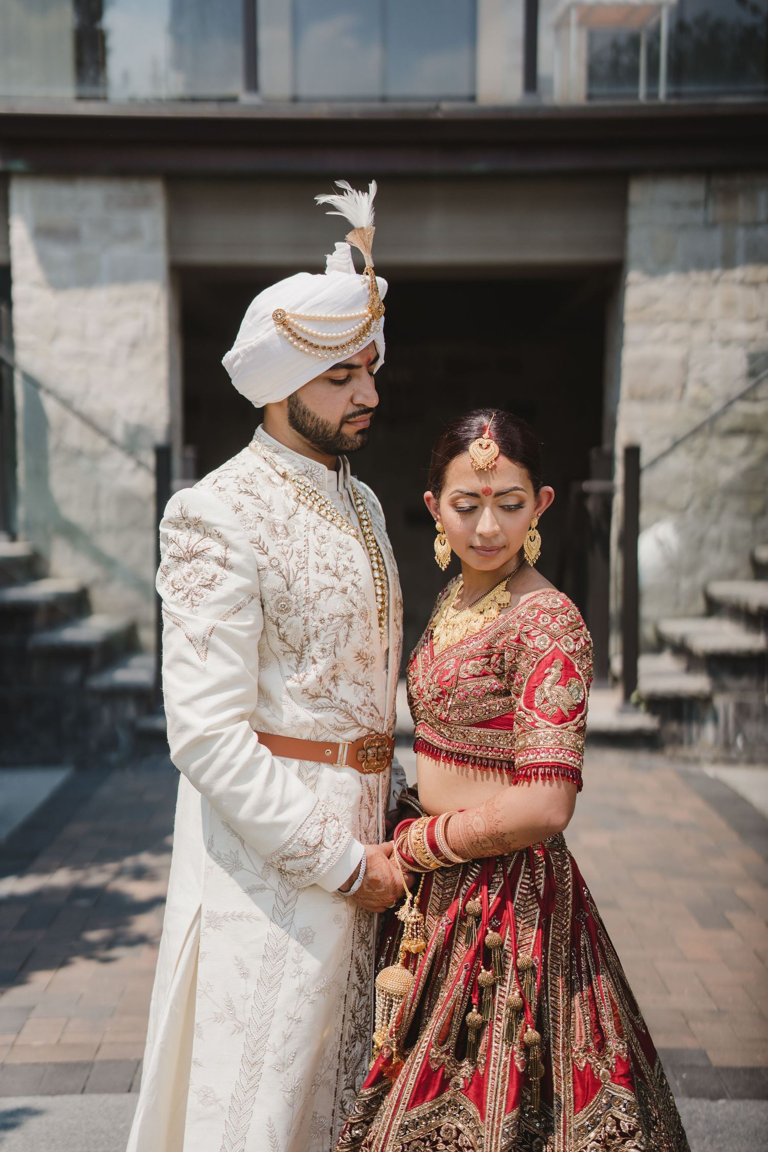 A couple in traditional South Asian wedding attire, with the bride in red lehenga and groom in white sherwani, pose together.