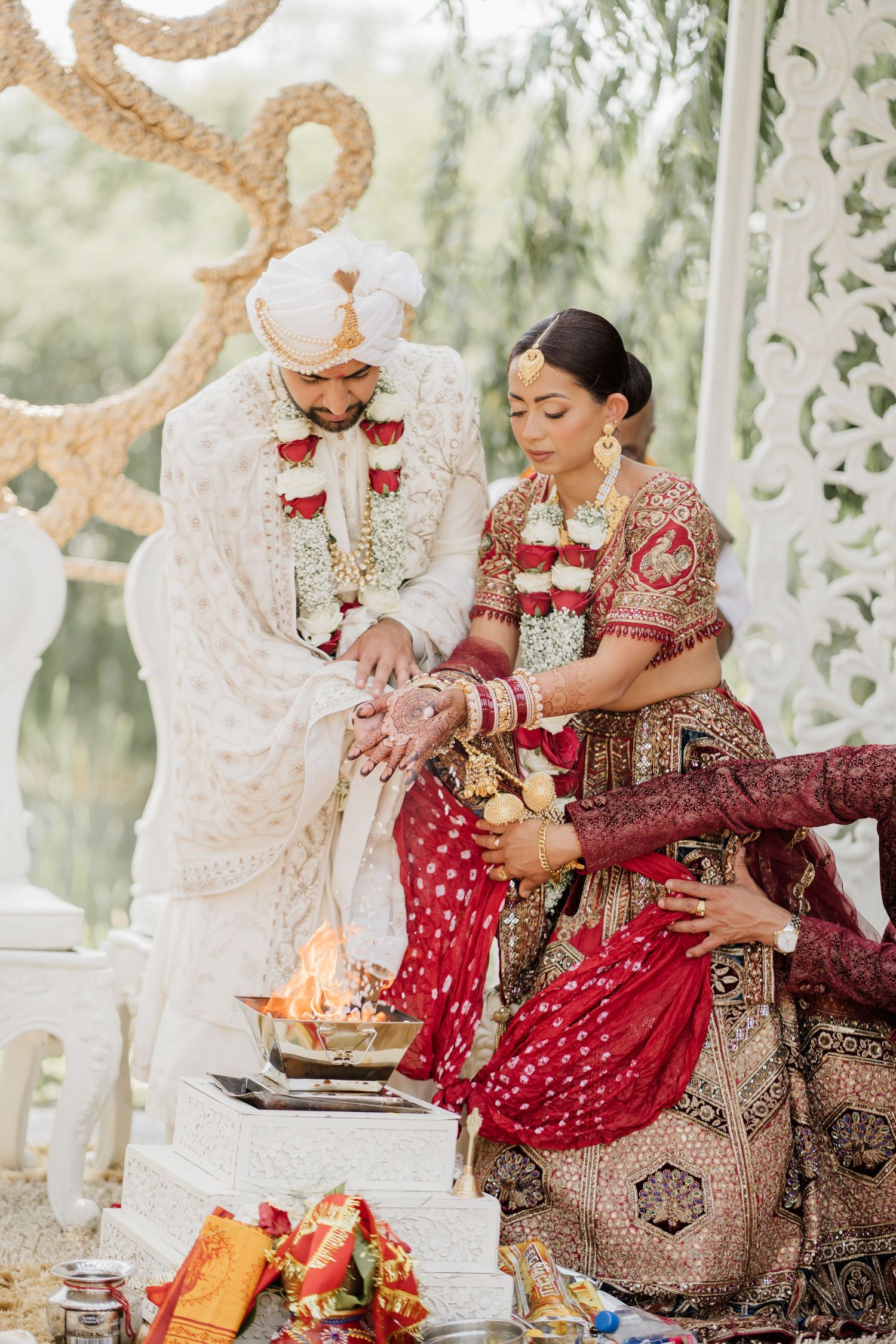 A traditional Indian wedding ceremony shows a couple performing sacred rituals with red and white decor elements.