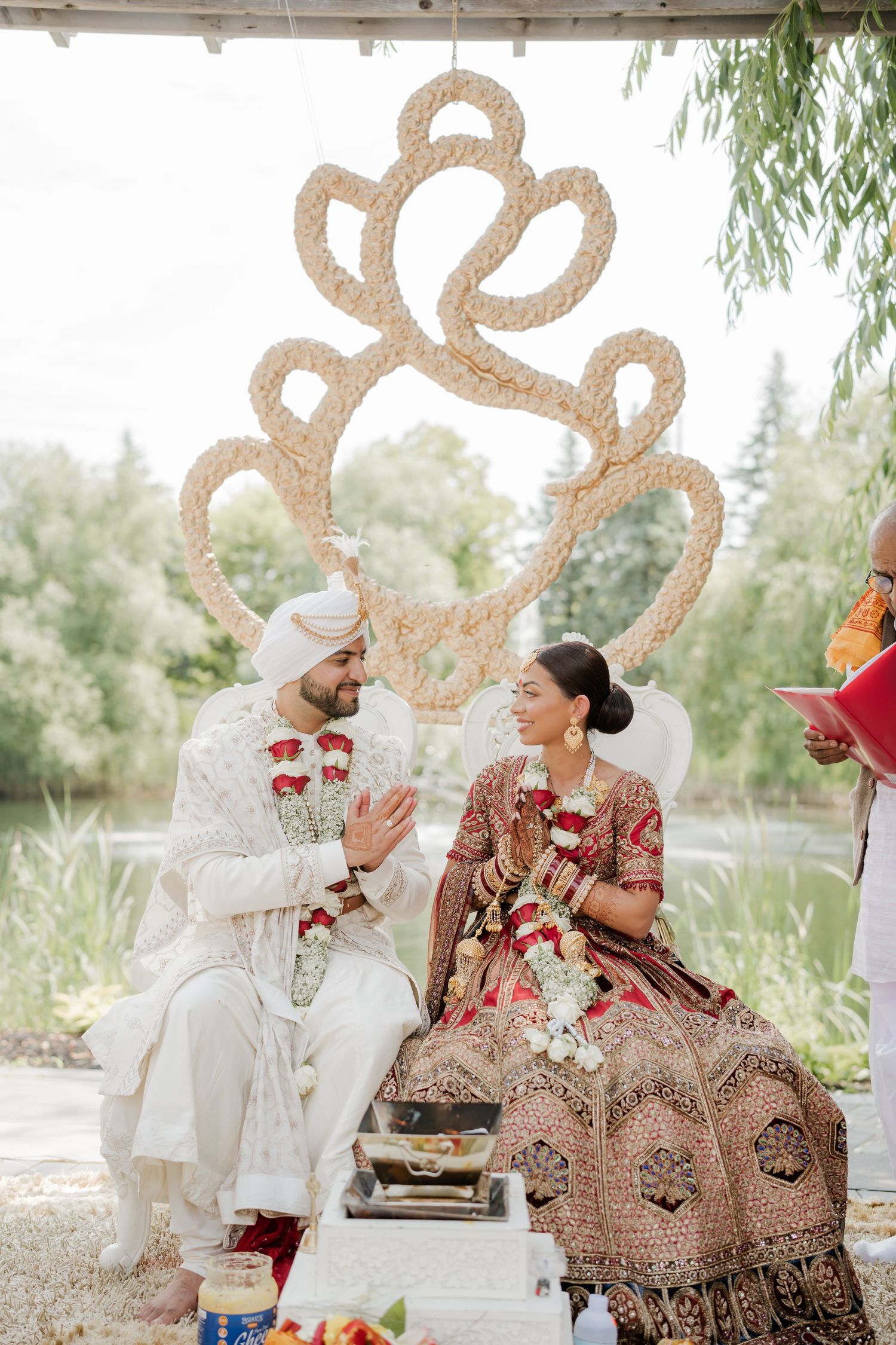 Traditional Indian wedding ceremony with couple seated under ornate decor and willow trees in outdoor garden setting.