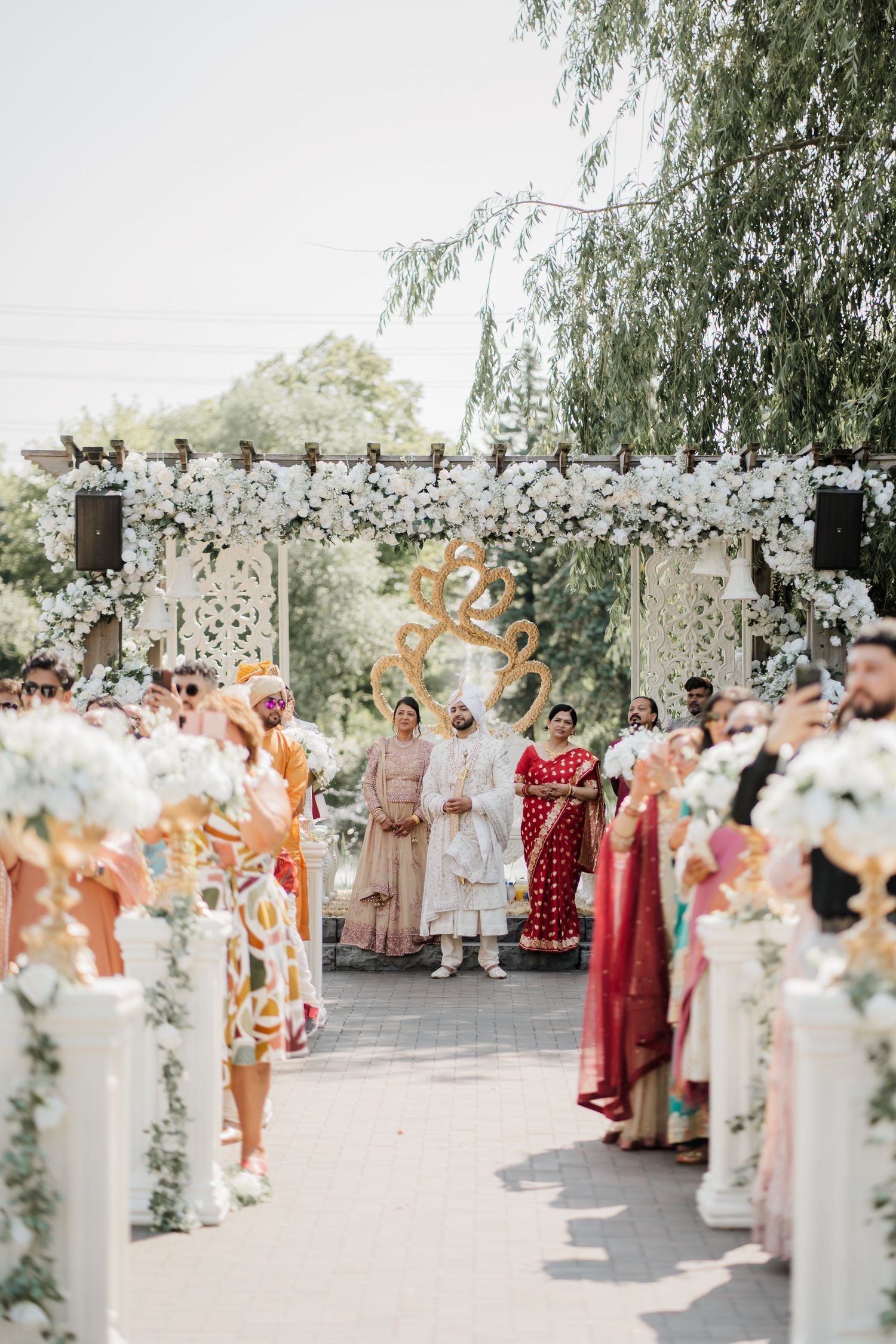 Wedding guests line up in formal attire under willow trees with floral decorations and white columns at an outdoor ceremony.
