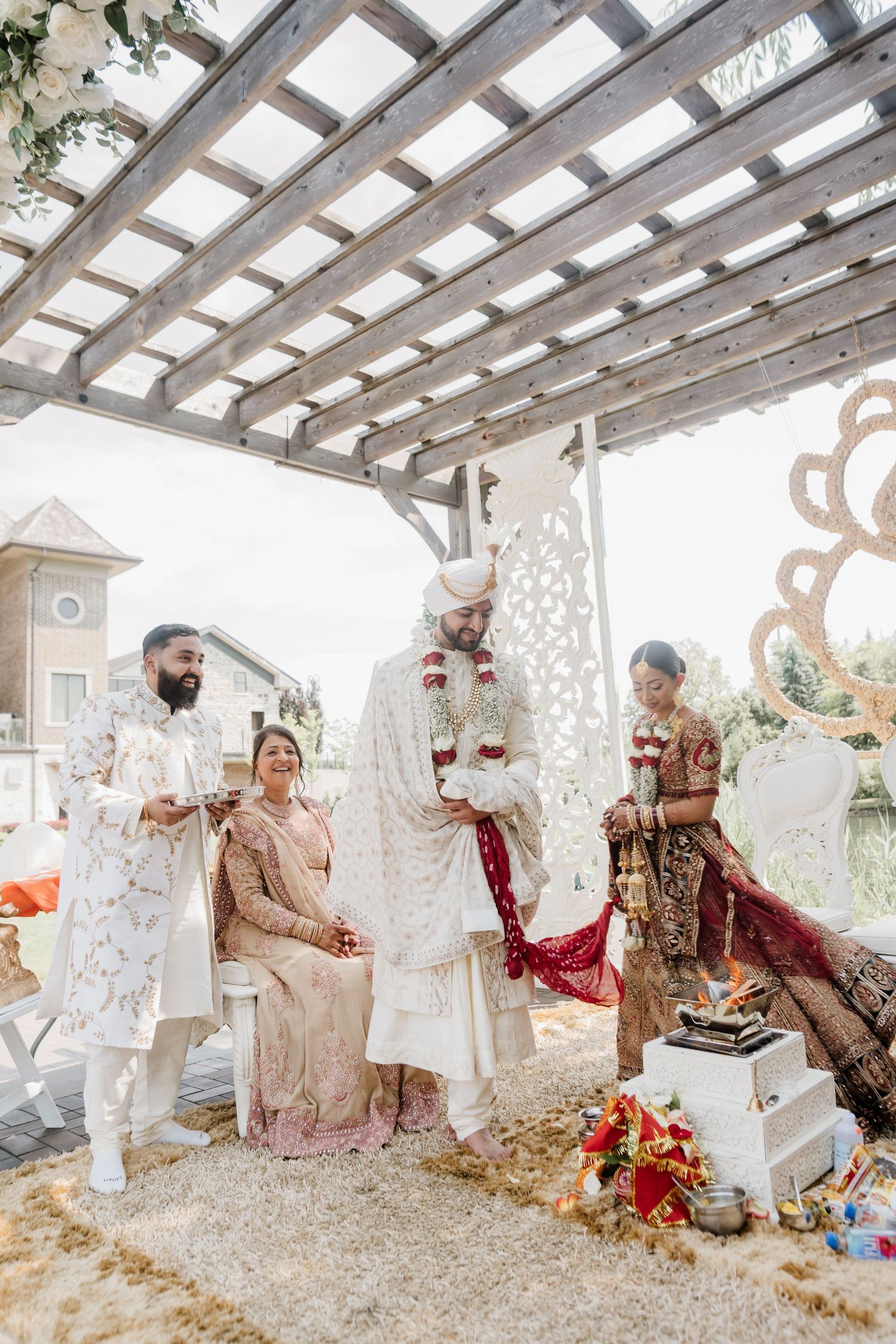 Couple in traditional South Asian wedding attire sits under a white pergola decorated with floral arrangements.