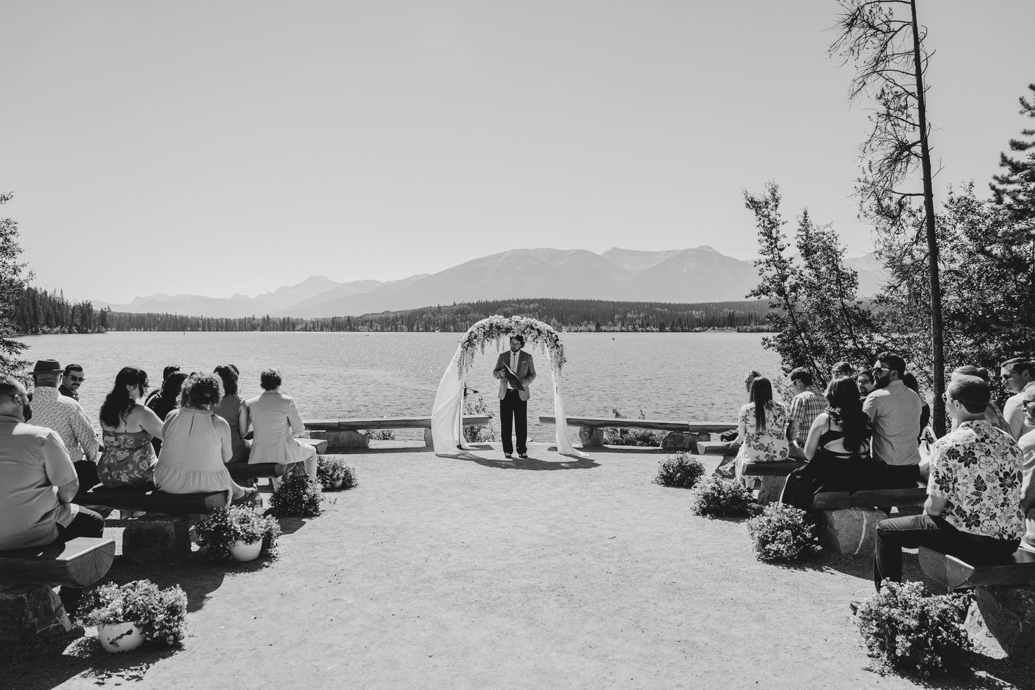 A panoramic view of a lakeside wedding ceremony with mountains and evergreen trees in the background.