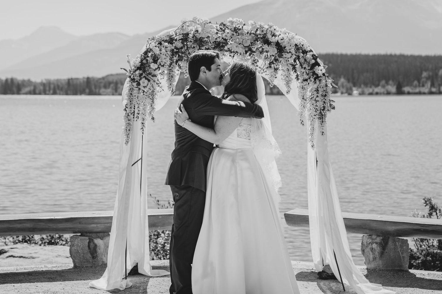 A romantic outdoor wedding ceremony takes place under a floral arch overlooking a serene lake.