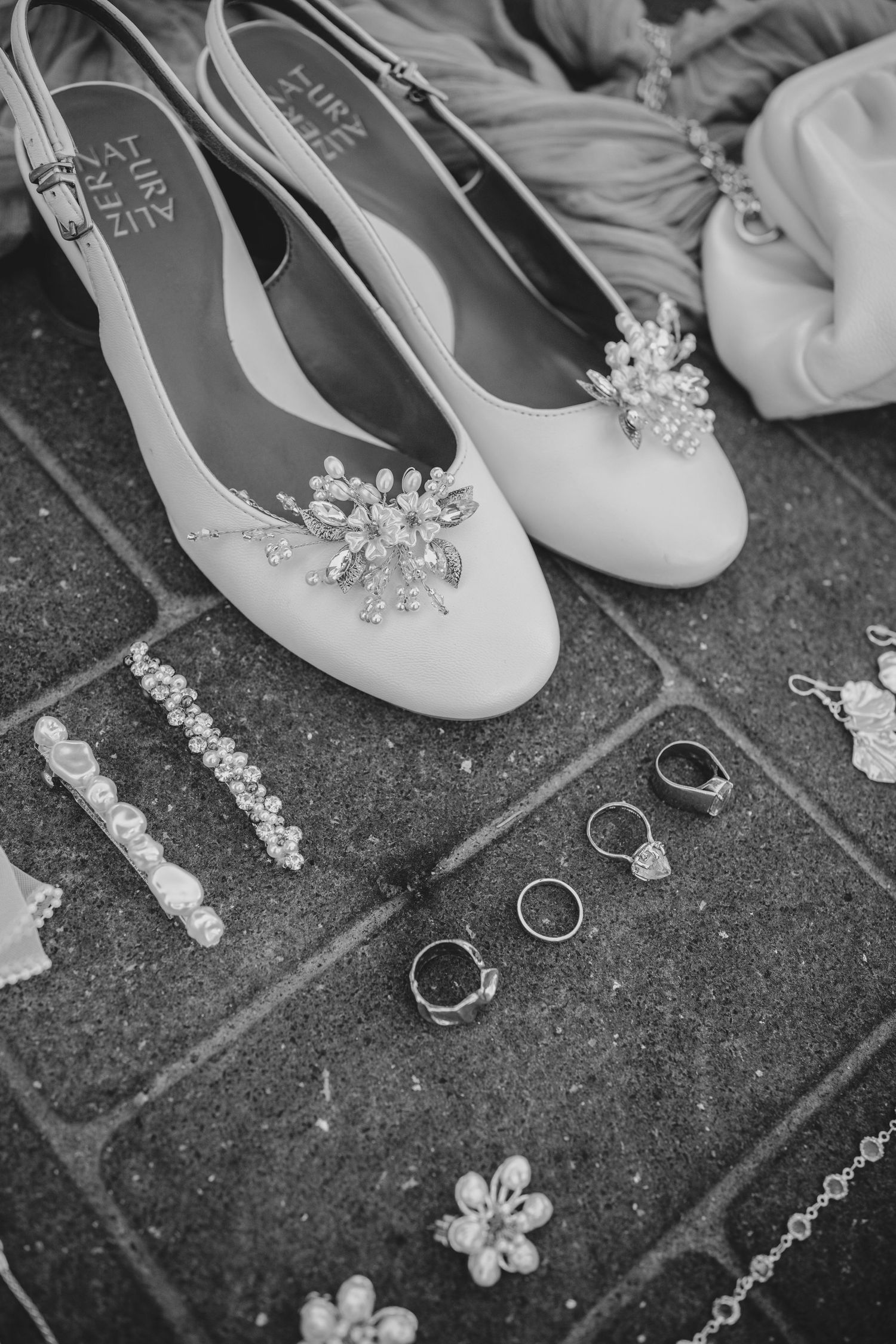 Wedding accessories arranged on stone surface including white floral shoes, rings, and delicate jewelry pieces.