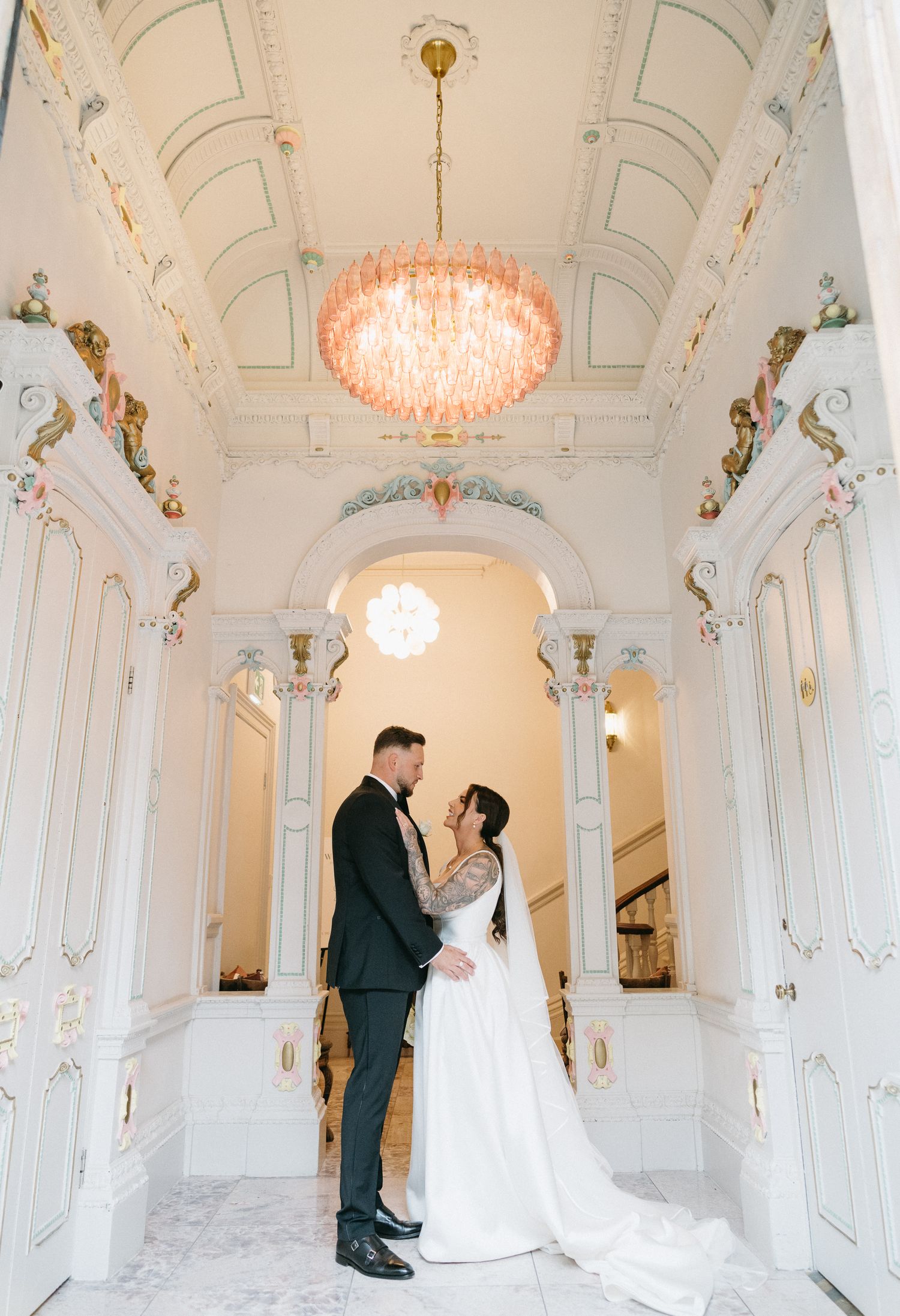Elegant wedding couple poses in ornate white baroque hallway with crystal chandeliers and decorative architectural details.