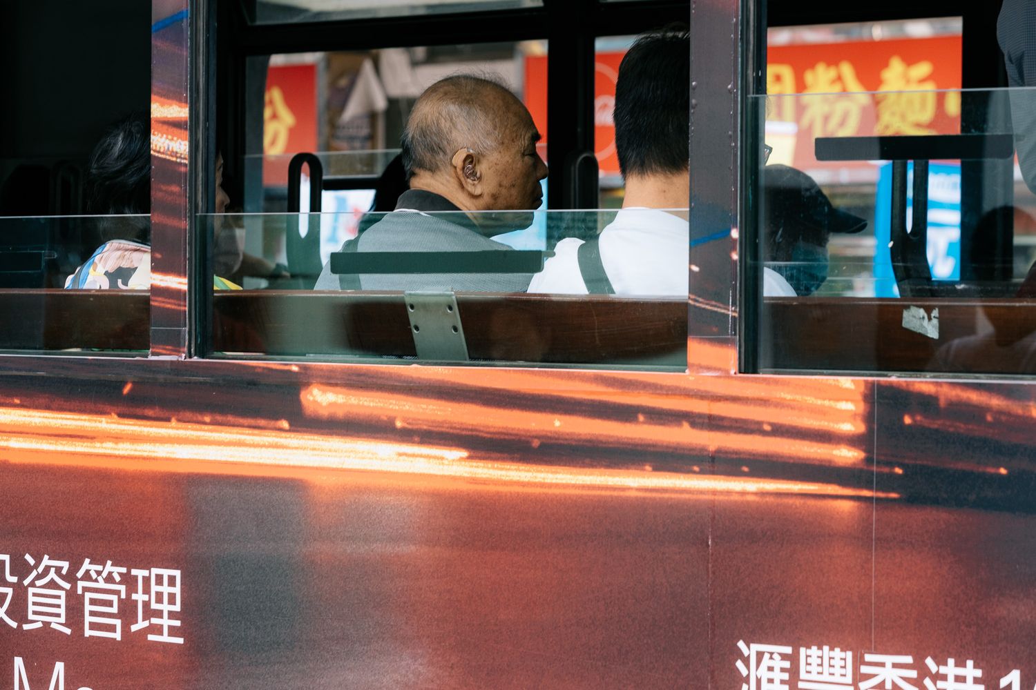 Elderly passengers seen through the reflective window of a Hong Kong bus with Chinese text signage.