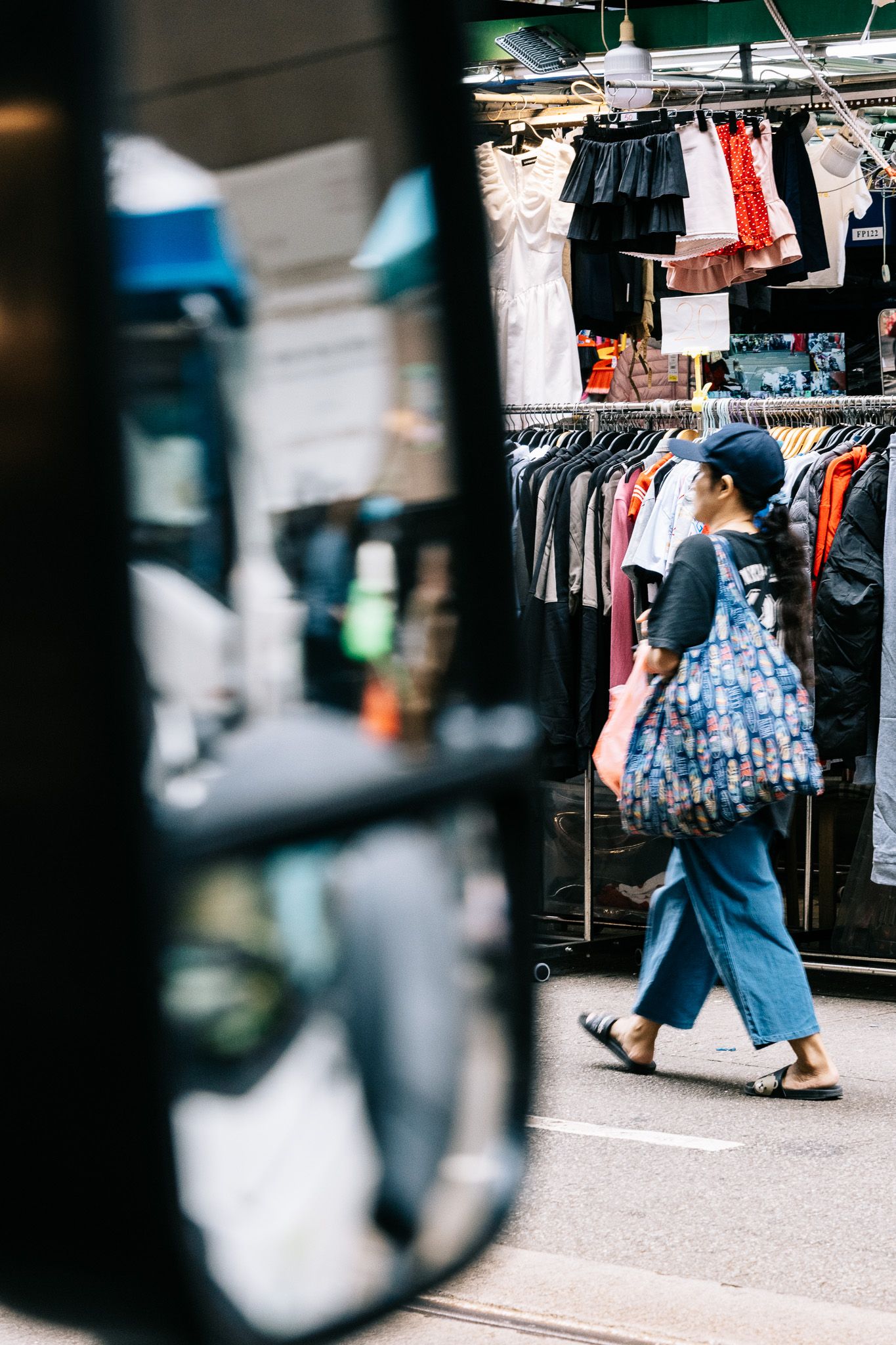Woman with large floral bag walking past outdoor clothing racks on a busy Hong Kong street market.