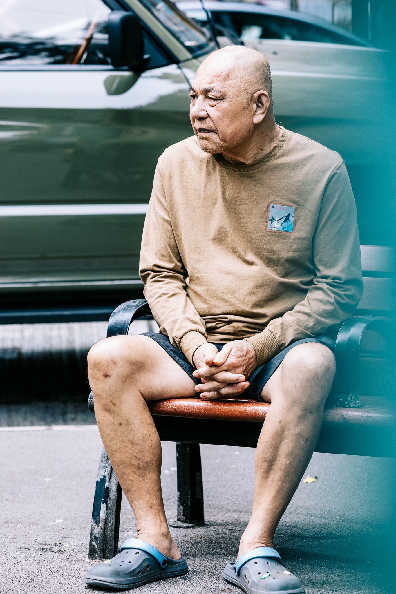 Elderly Asian man sitting on a bench outdoors, wearing a beige sweater and shorts with blue Crocs sandals.