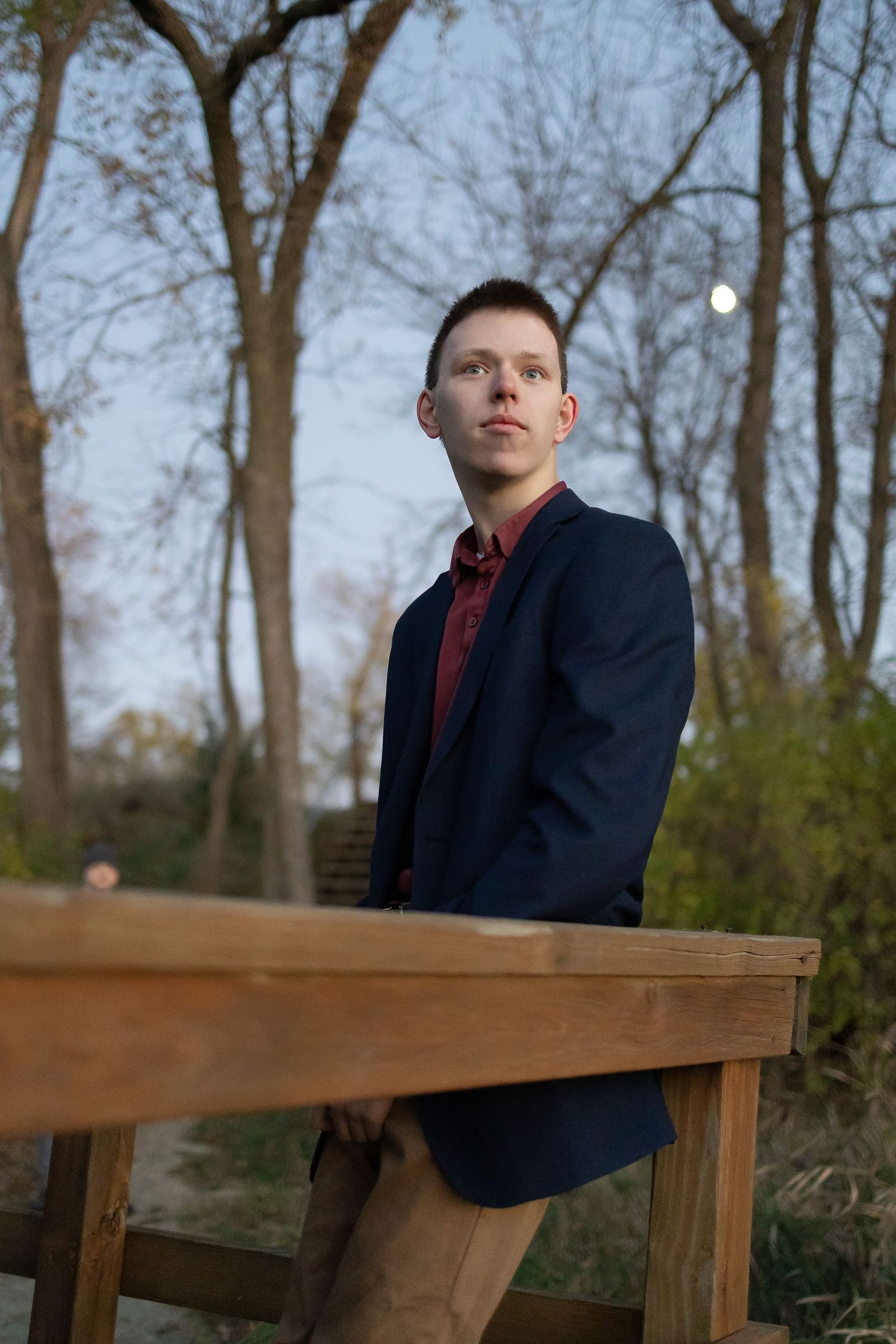 Sioux City, Iowa photographer. Young man in navy blazer and red shirt leans on wooden railing outdoors at dusk.