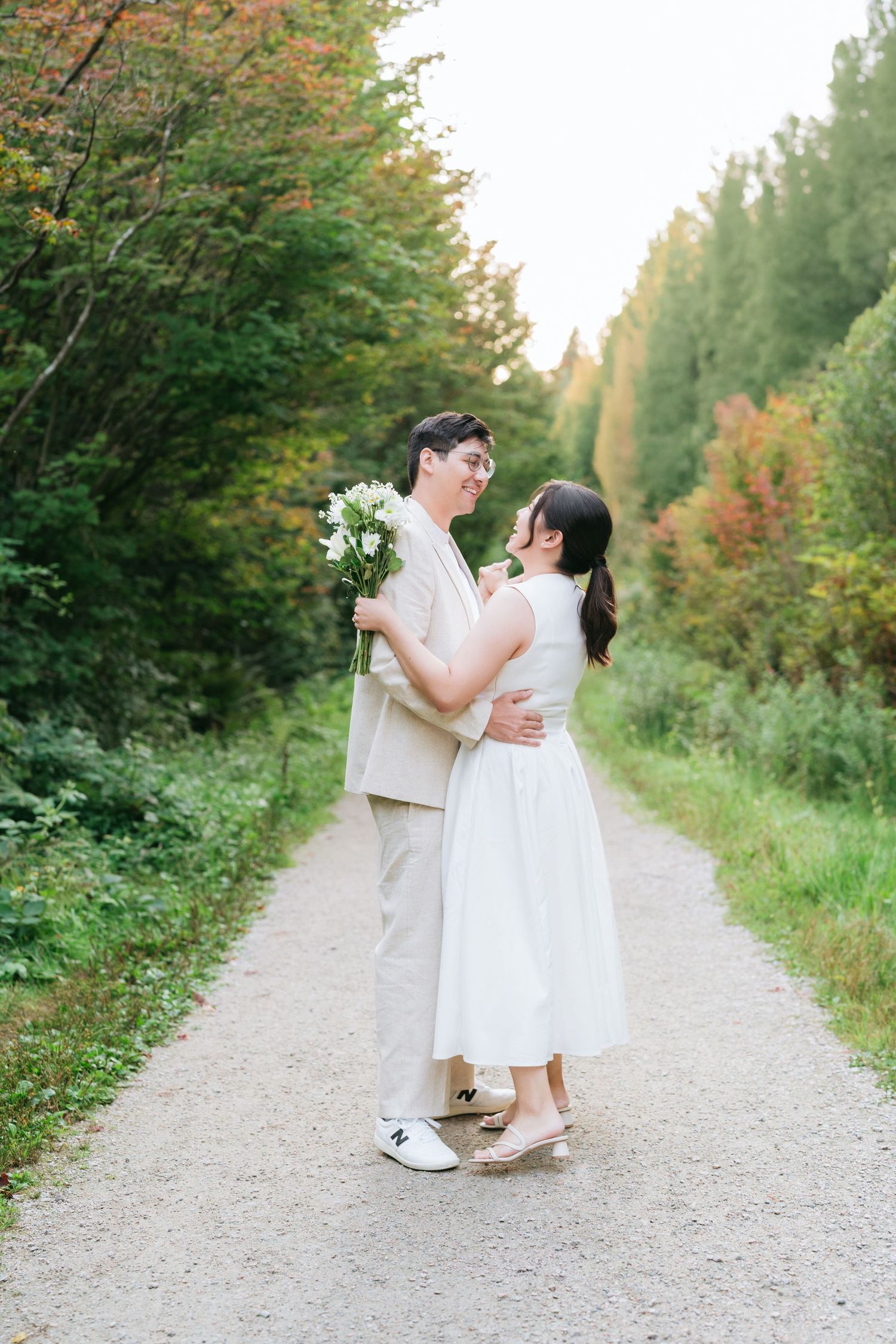 A couple in light colored formal attire dances playfully along a tree-lined gravel path during a romantic photoshoot.