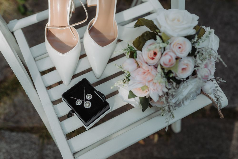 White bridal heels, delicate pink roses, and a black ring box arranged on a white wooden chair for wedding detail photography.