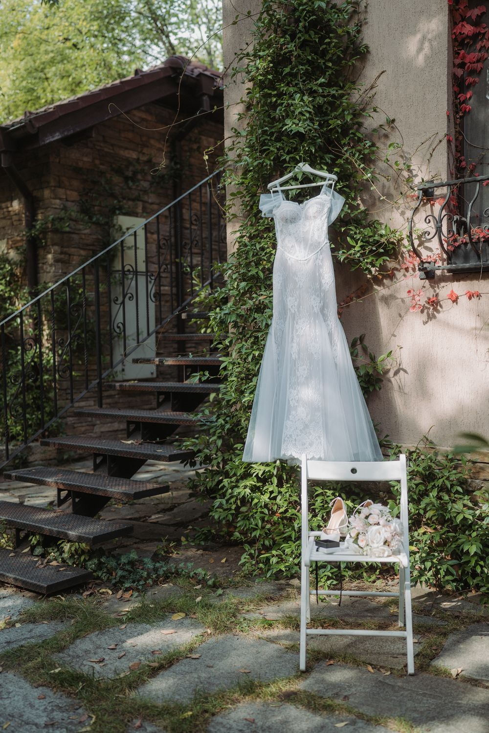 White wedding dress hanging on a ladder chair against a rustic stone wall with climbing vines and wooden stairs.