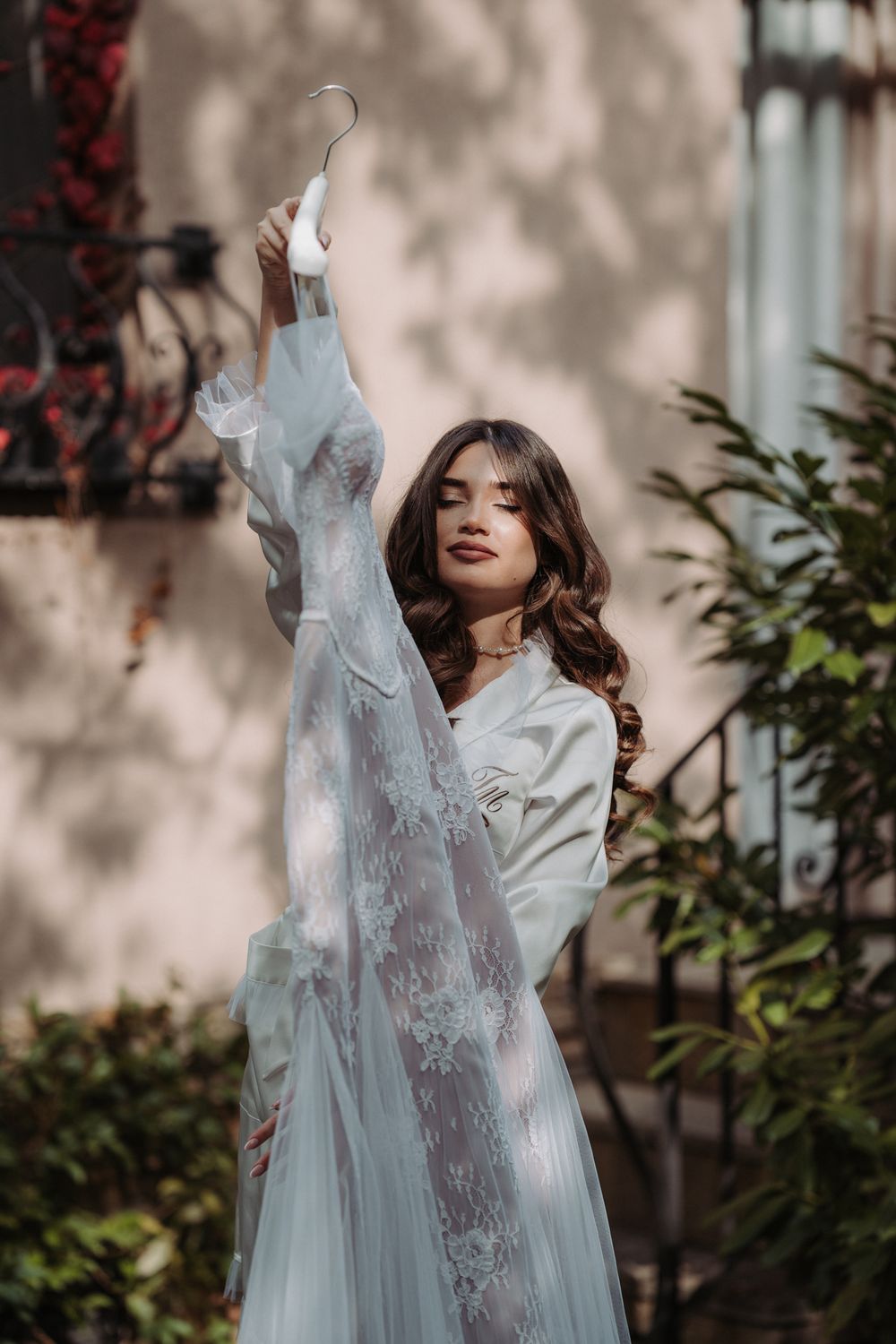 A woman in a flowing white lace gown poses gracefully in a garden setting with dappled sunlight.