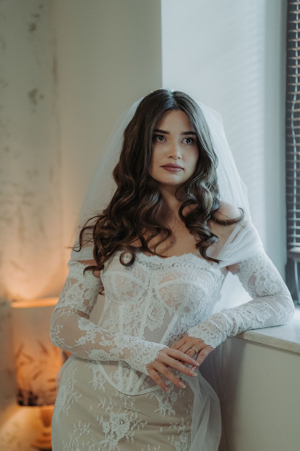A bride in a long-sleeved lace wedding dress and veil poses near a window with soft lighting.