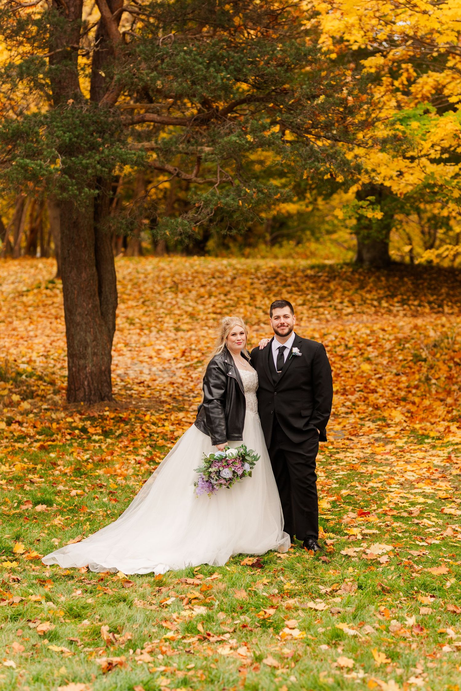 A couple poses for wedding photos in a park with vibrant autumn foliage and fallen leaves on the ground.