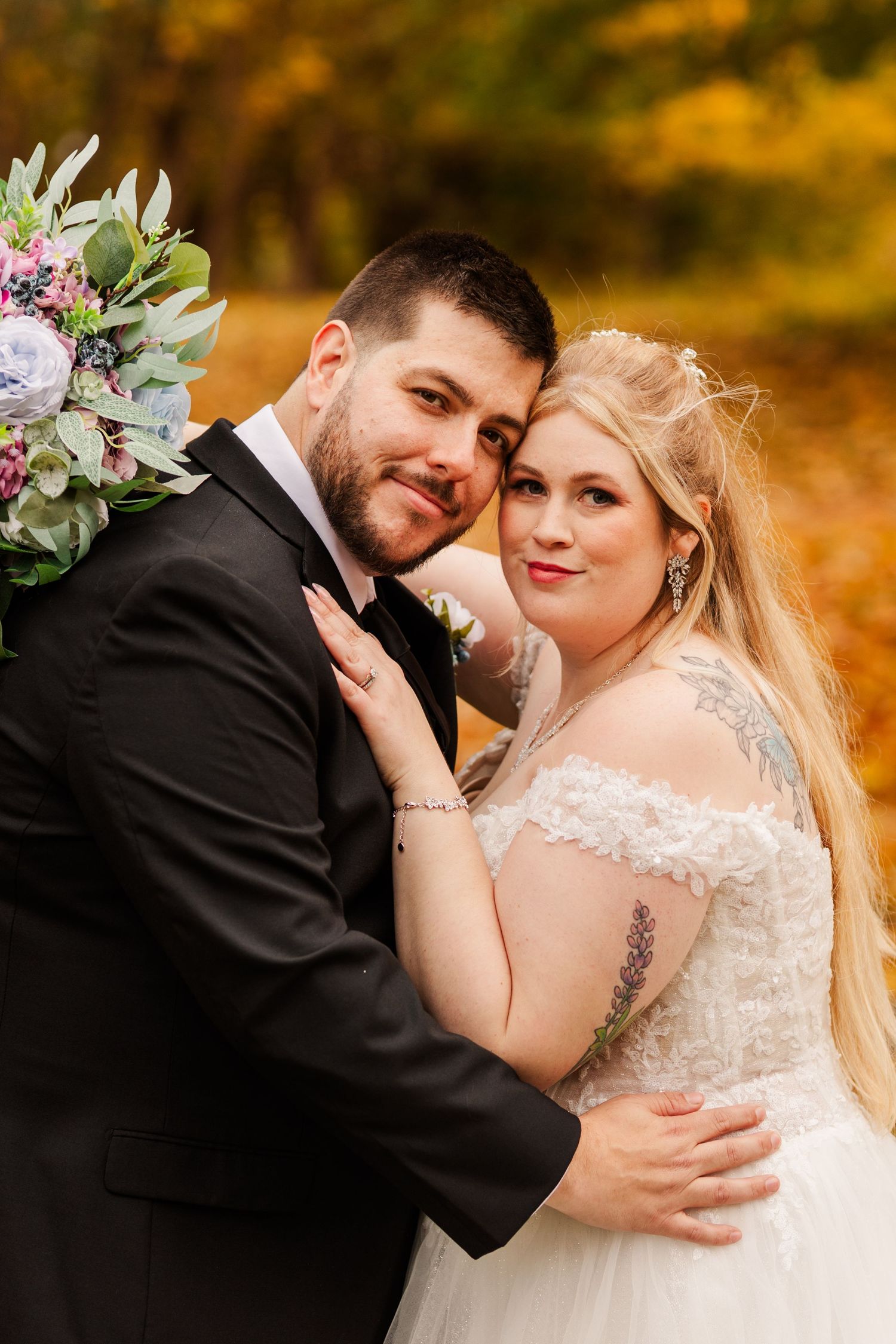 A romantic wedding portrait of a couple embracing outdoors with autumn foliage and a beautiful floral bouquet.