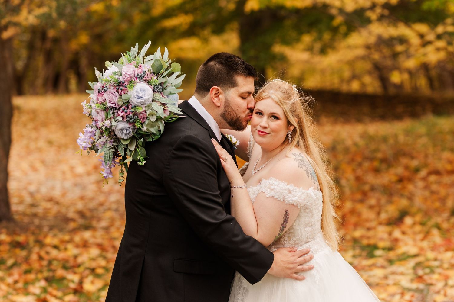 Romantic autumn wedding couple embraces surrounded by vibrant orange fall foliage in an outdoor setting.
